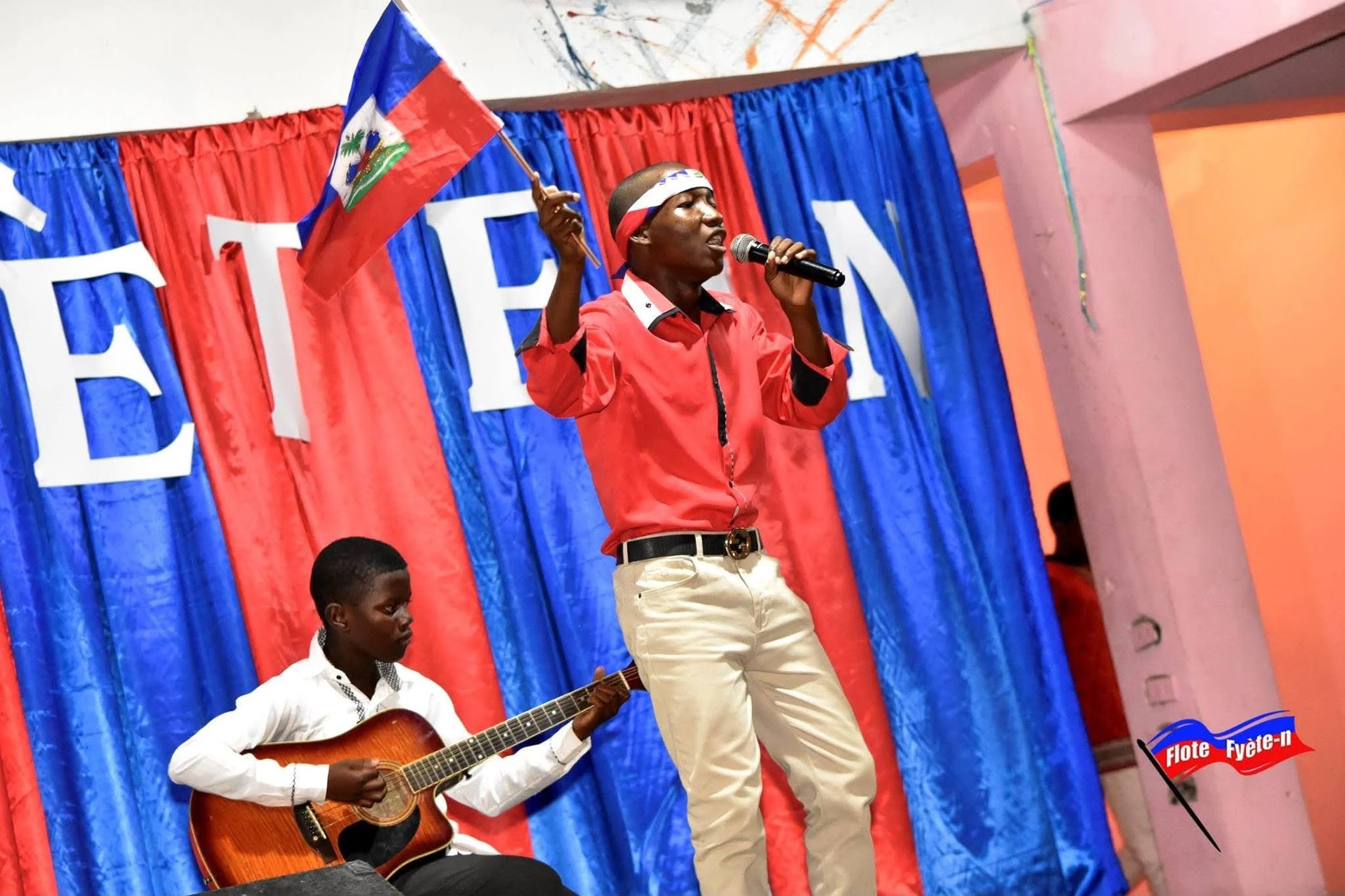 Un homme chante en tenant un drapeau haïtien, tandis qu'un homme joue de la guitare à côté de lui. Le fond est composé de rideaux rouges et bleus avec un décor de fête.