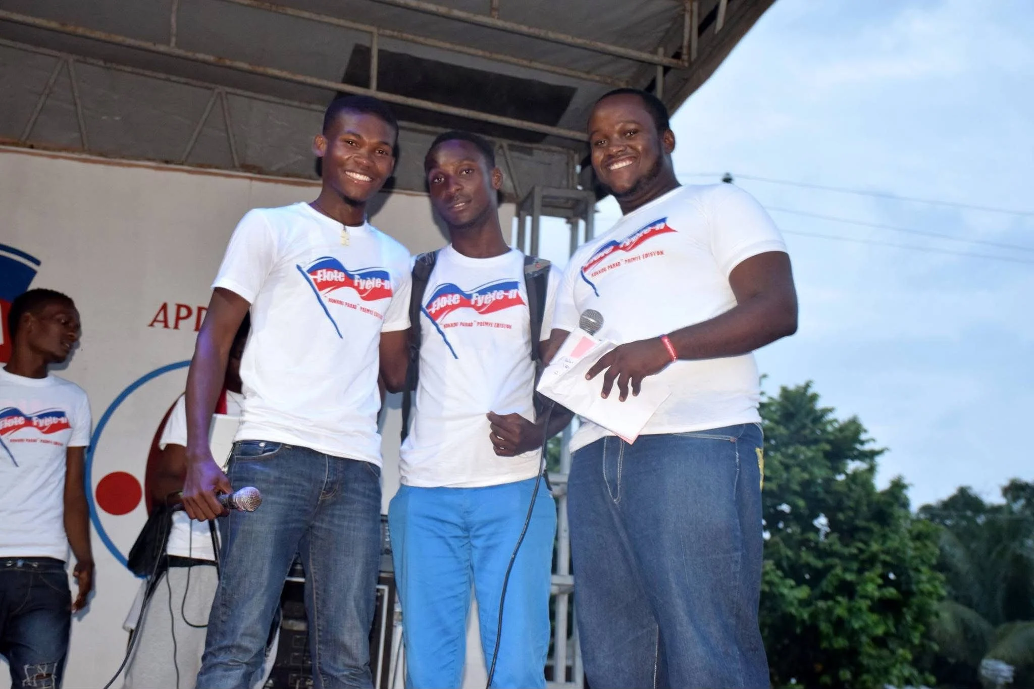 Trois hommes souriants portant des T-shirts avec un logo, se tenant sur une scène lors d'un événement, avec un micro dans une main de l'un d'eux.