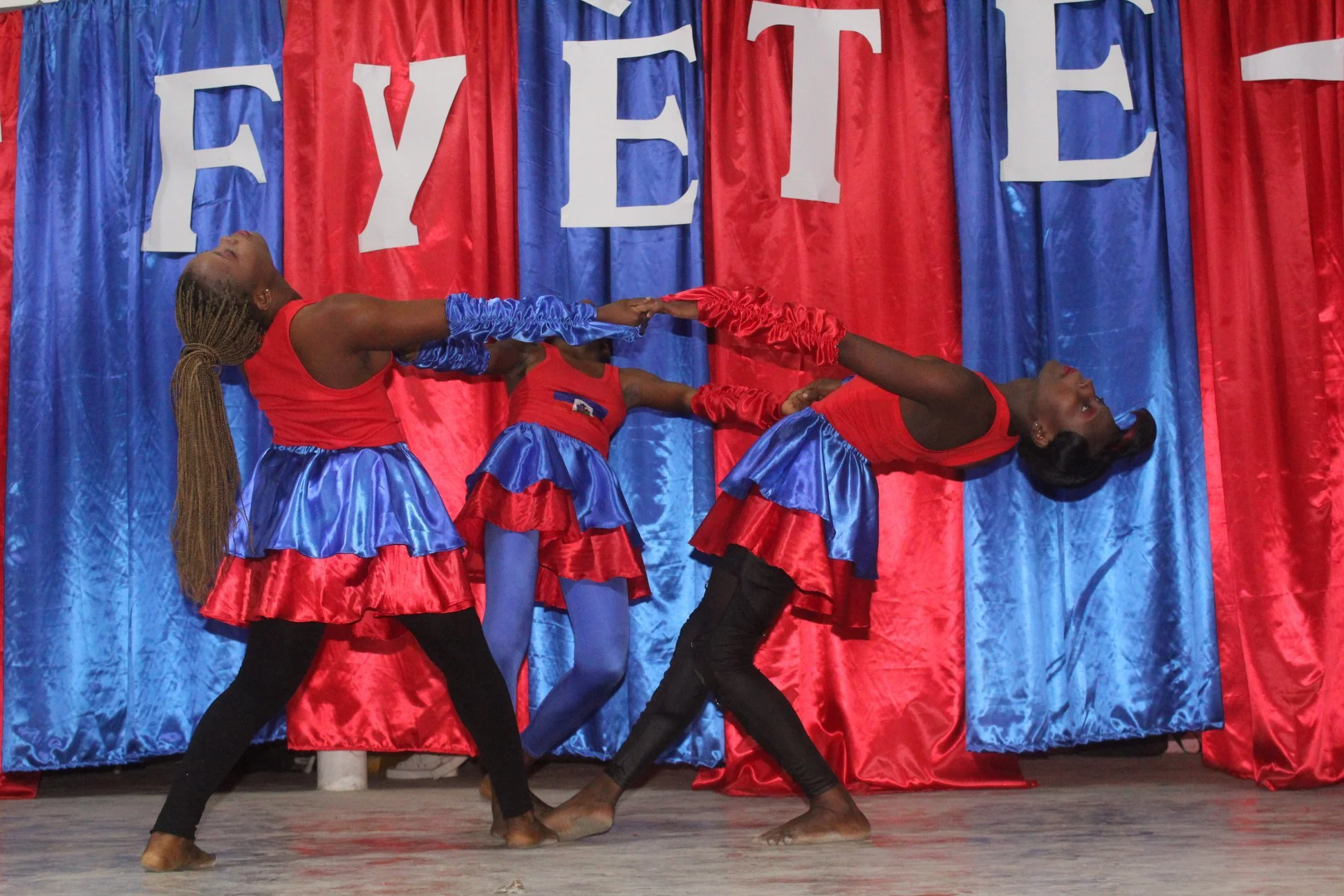 Trois jeunes filles en costumes bleus et rouges dans une performance de danse sur scène avec un drap de fond rouge et bleu avec le mot 'FYE'.