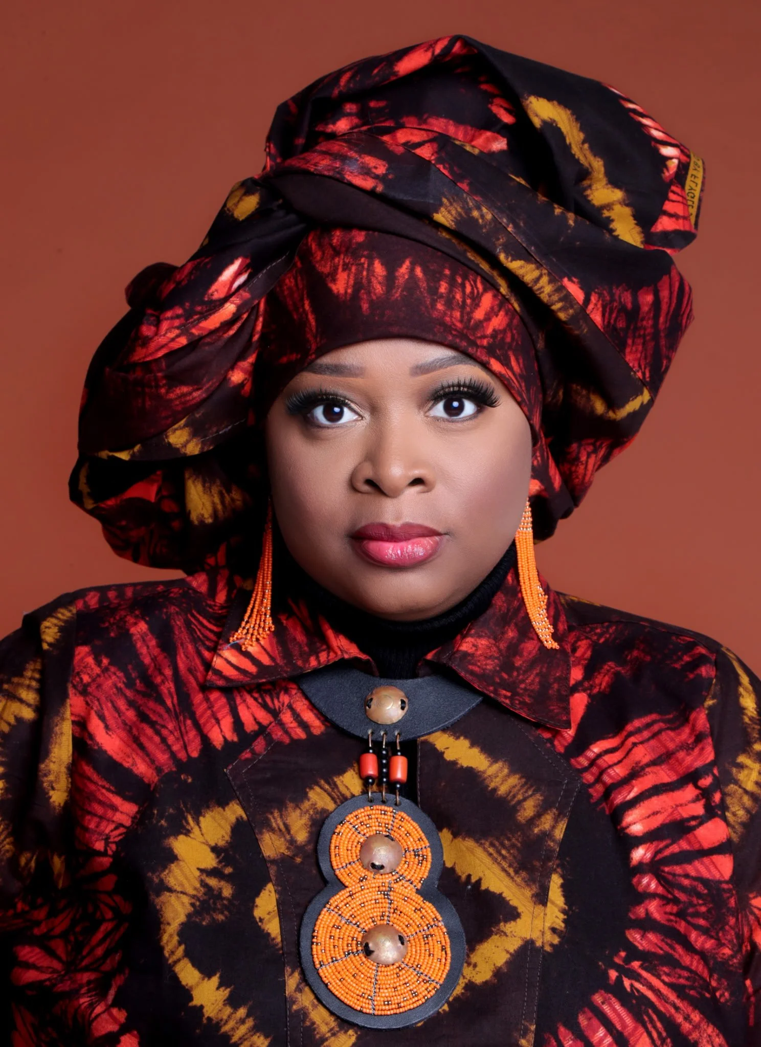 Une femme portant un vêtement traditionnel africain rouge et noir avec un grand turban assorti, des boucles d'oreilles orange et un collier avec un pendentif rond brodé de perles orange.