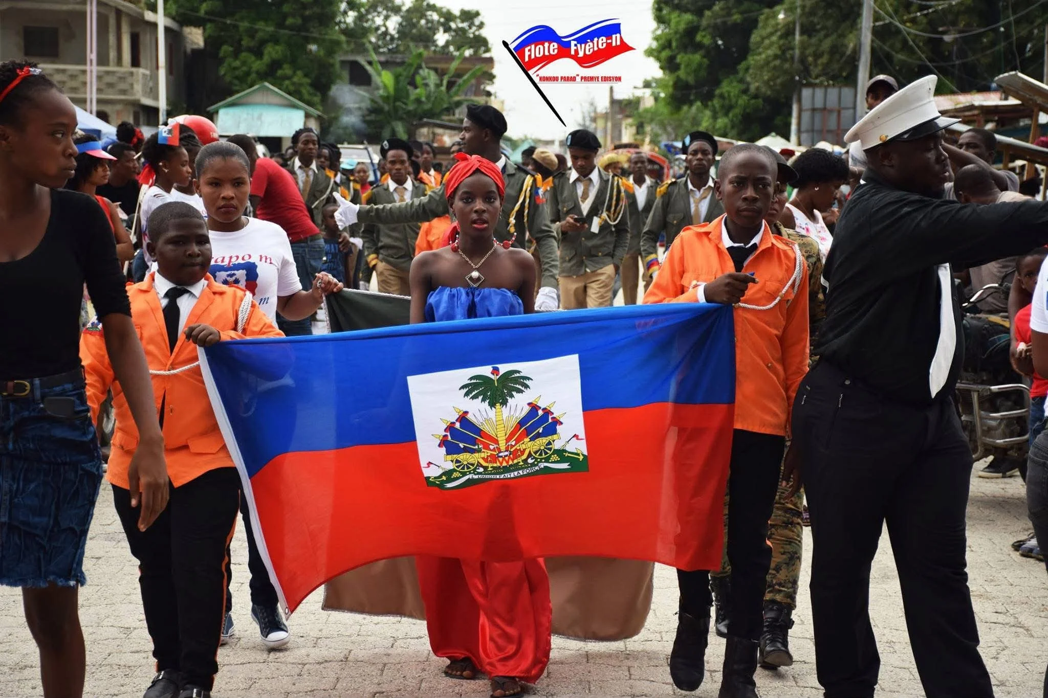 Une parade dans une rue en Haïti, avec des jeunes et des adultes, certains portant des uniformes et brandissant un drapeau d'Haïti.