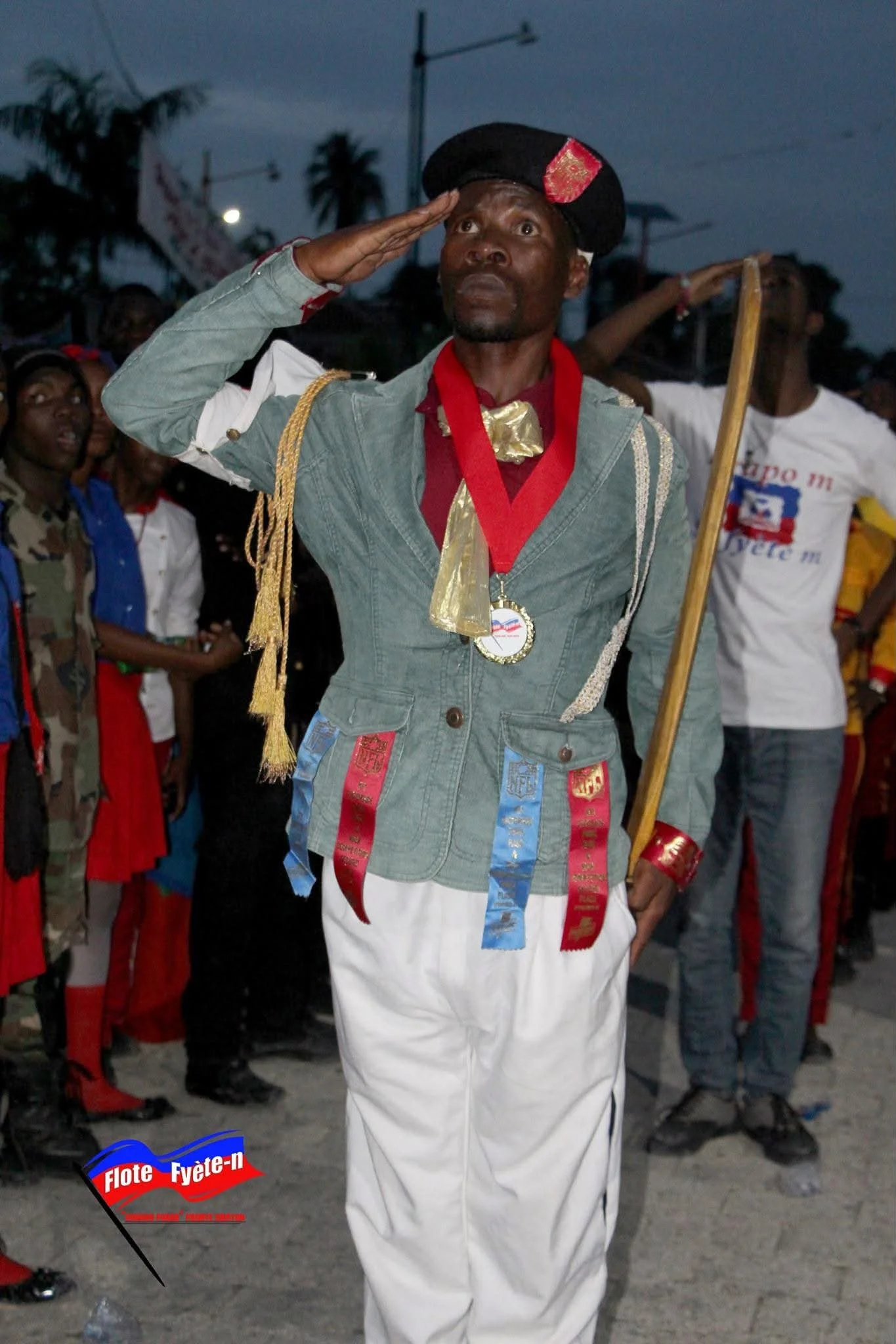 Un homme en uniforme de marin, portant un chapeau noir orné de rouge, fait un salut militaire avec sa main droite. Il porte également une médaille, une écharpe rouge, et des décorations colorées sur sa veste. Il se tient dans une foule lors d'une cér