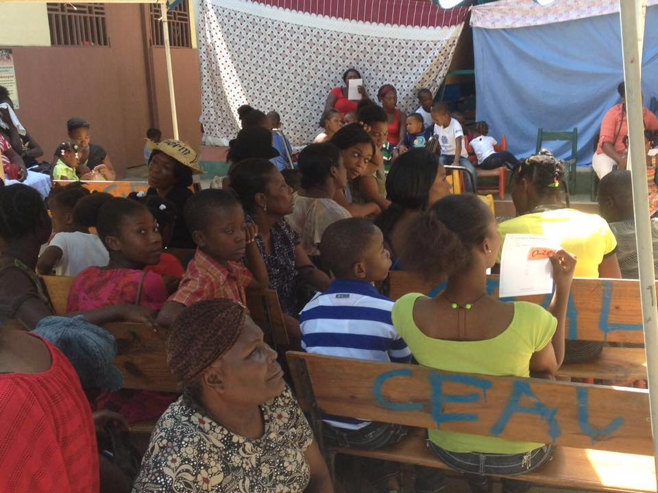 Un groupe de personnes, principalement des femmes, assises sur des bancs lors d'une réunion ou d'un événement en plein air. Certains prennent des notes ou utilisent des téléphones, sous une tente ou un abri."