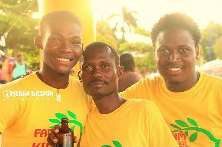 Trois jeunes hommes portant des t-shirts jaunes avec un logo vert, souriant et posant pour la photo lors d'un événement en plein air, avec un fond ensoleillé et des arbres.