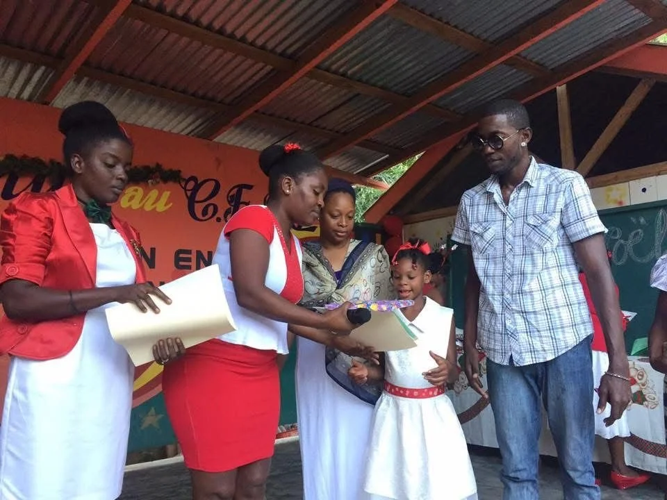 Une cérémonie scolaire où une jeune fille reçoit un certificat ou un prix d'une femme en uniforme, entourée de deux autres femmes et d'un homme. La scène se déroule sous un toit en bois lors d'une célébration ou remise de récompenses.