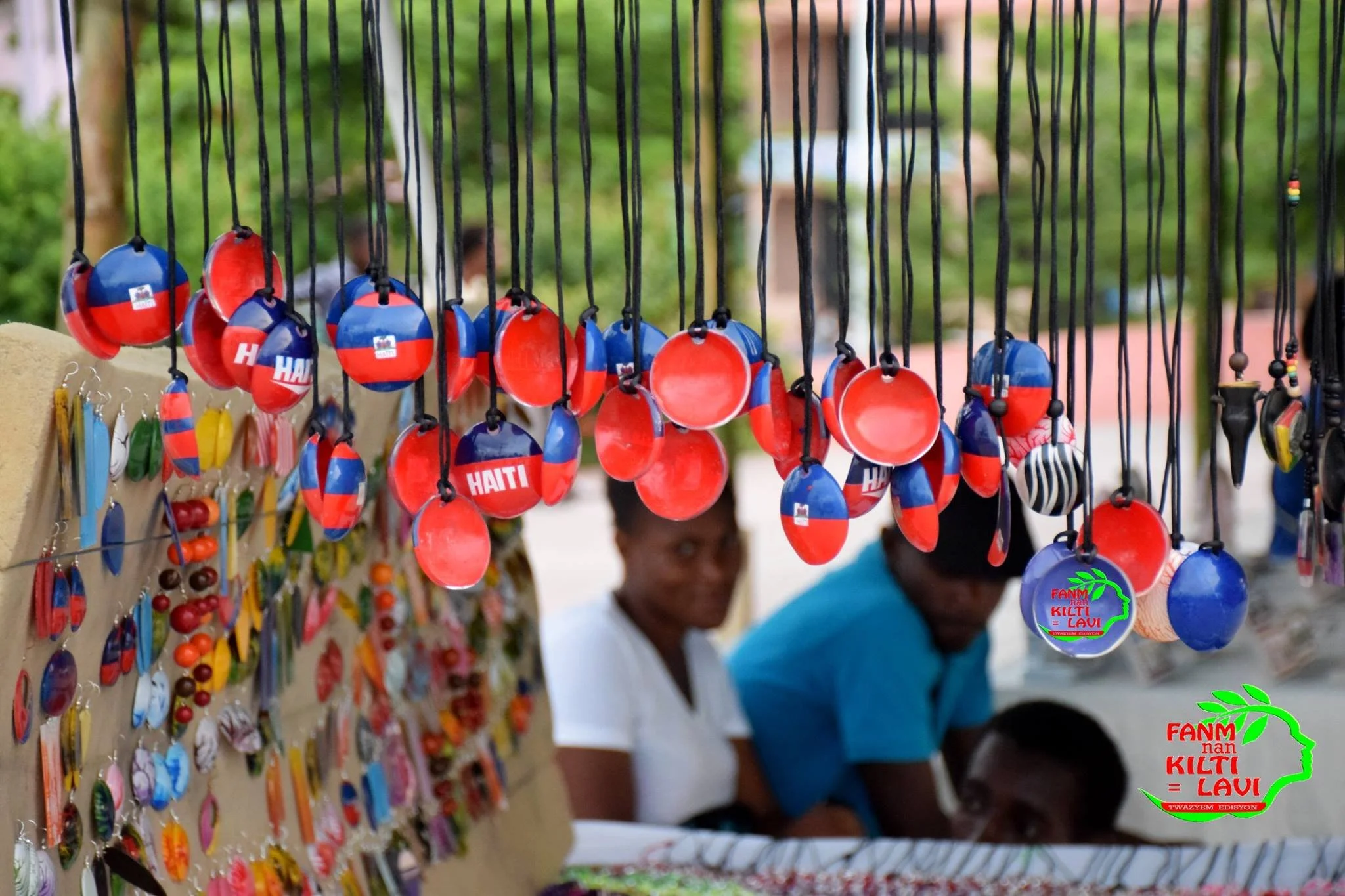 Épingles et souvenirs colorés, avec le texte "FANMI nan KILTI = LAWI" et une carte d'Haïti en arrière-plan.