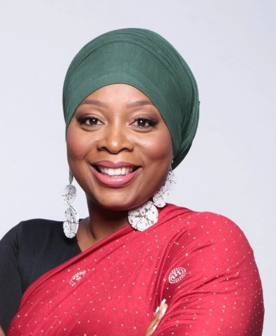 Femme souriante portant un turban vert, une robe rouge avec des motifs, et de grandes boucles d'oreilles en argent.