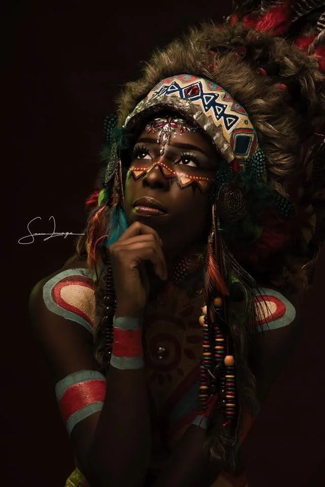 Une femme portant un costume traditionnel africain avec une coiffe ornée de plumes et de bijoux, peinte avec des motifs colorés sur la peau, en pose réfléchie.