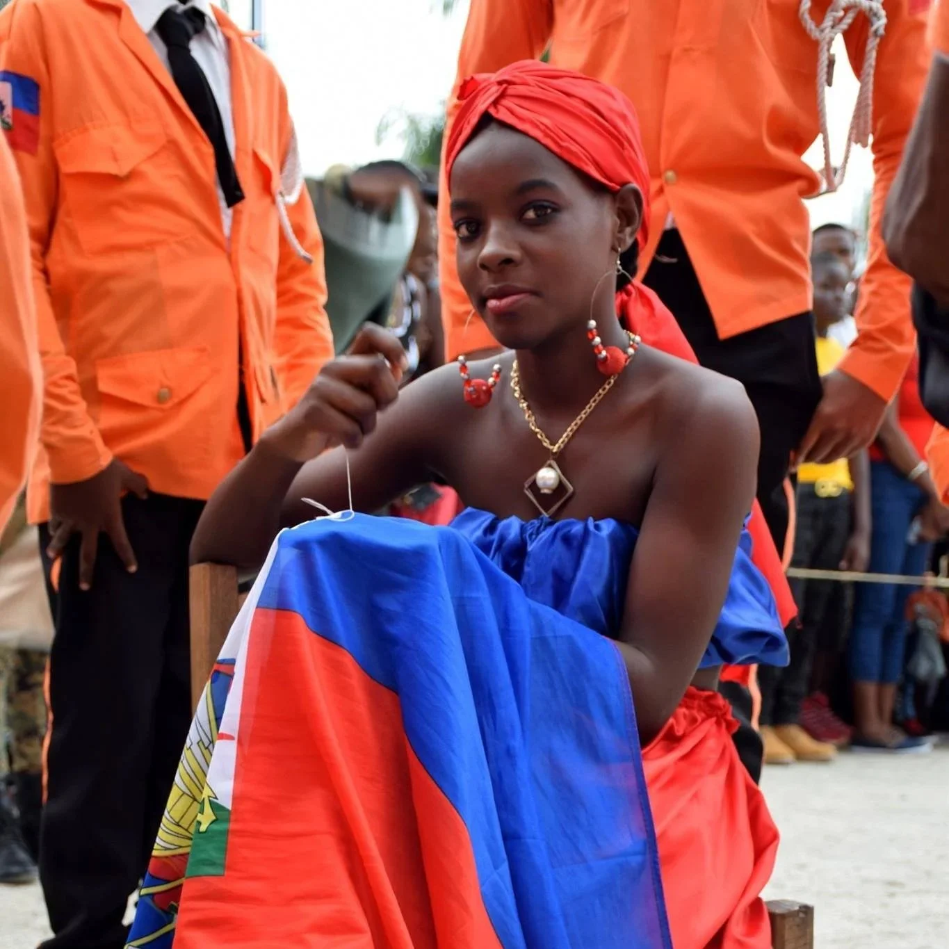 Une jeune femme portant un tissu aux couleurs du drapeau d'Haïti, ornée de bijoux, assise lors d'un événement, entourée de personnes en uniformes orange.