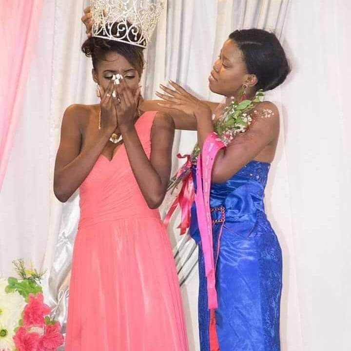 Deux femmes en robes de soirée lors d'une célébration, l'une porte une couronne et semble émue, l'autre lui donne un soutien avec un sourire.