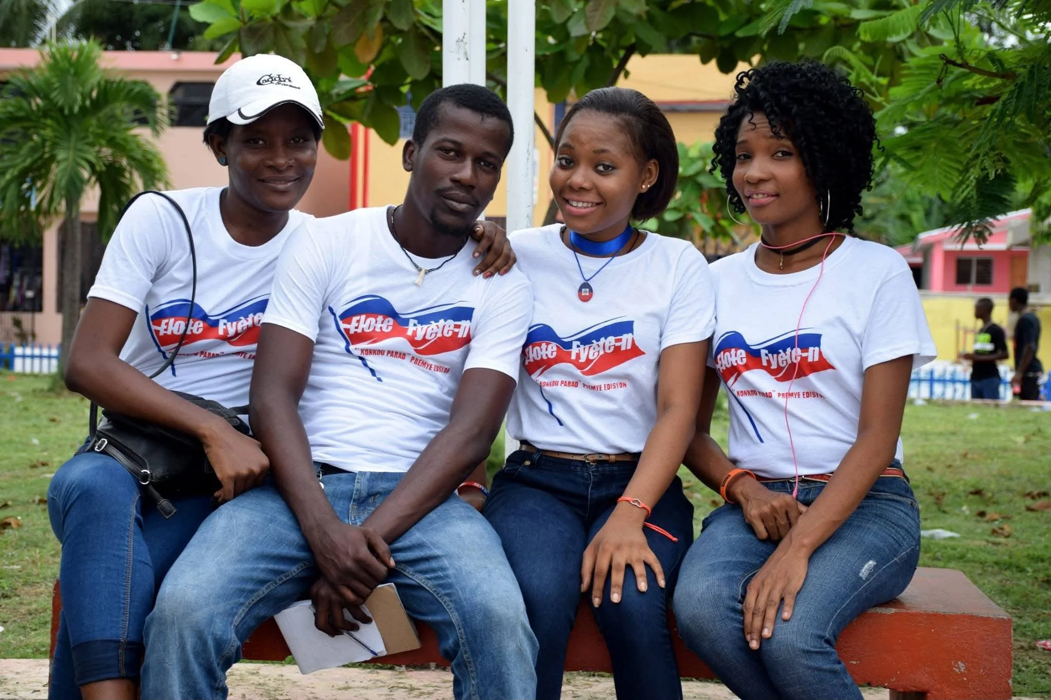 Groupe de quatre jeunes femmes et hommes assis sur un banc dans un parc, portant des t-shirts blancs avec un logo rouge, blanc et bleu, souriant et posant pour la photo.