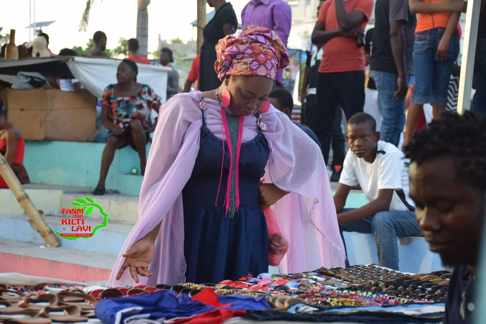 Une femme portant un foulard coloré, regardant des bijoux artisanaux à un marché en plein air, avec d'autres personnes en arrière-plan. Logo en bas à gauche avec texte "FANM NÀN KILTI LAWI".