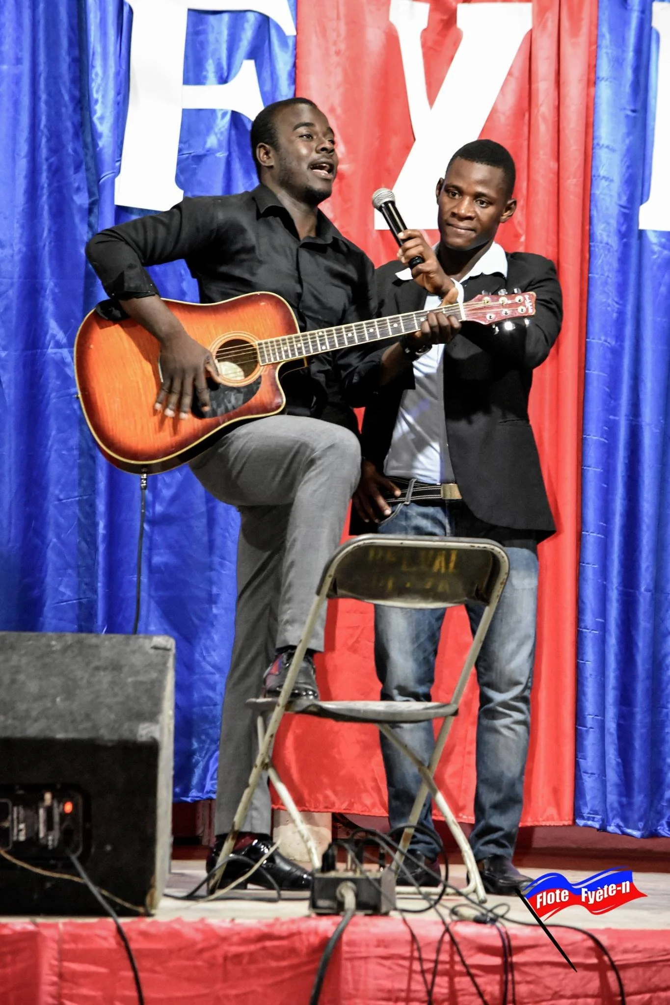 Deux hommes sur scène, un guitare acoustique, un microphone, un fond avec du tissu rouge et bleu, un drapeau avec le texte 'Fête-n'.