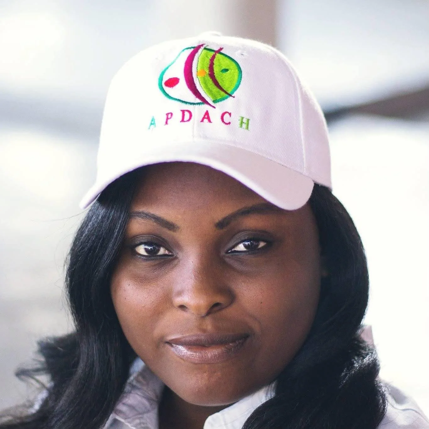 Une femme portant une casquette blanche avec un logo coloré et le texte APDACH. Elle a les cheveux noirs longs et un maquillage naturel.