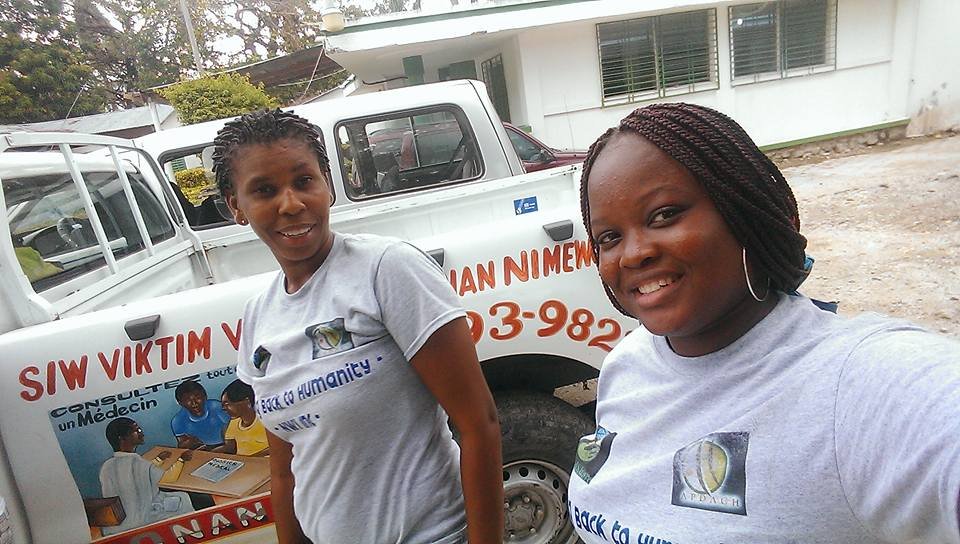 Deux femmes portent des t-shirts gris avec des inscriptions. Elles posent devant un véhicule blanc avec un logo et un texte en français sur le côté.