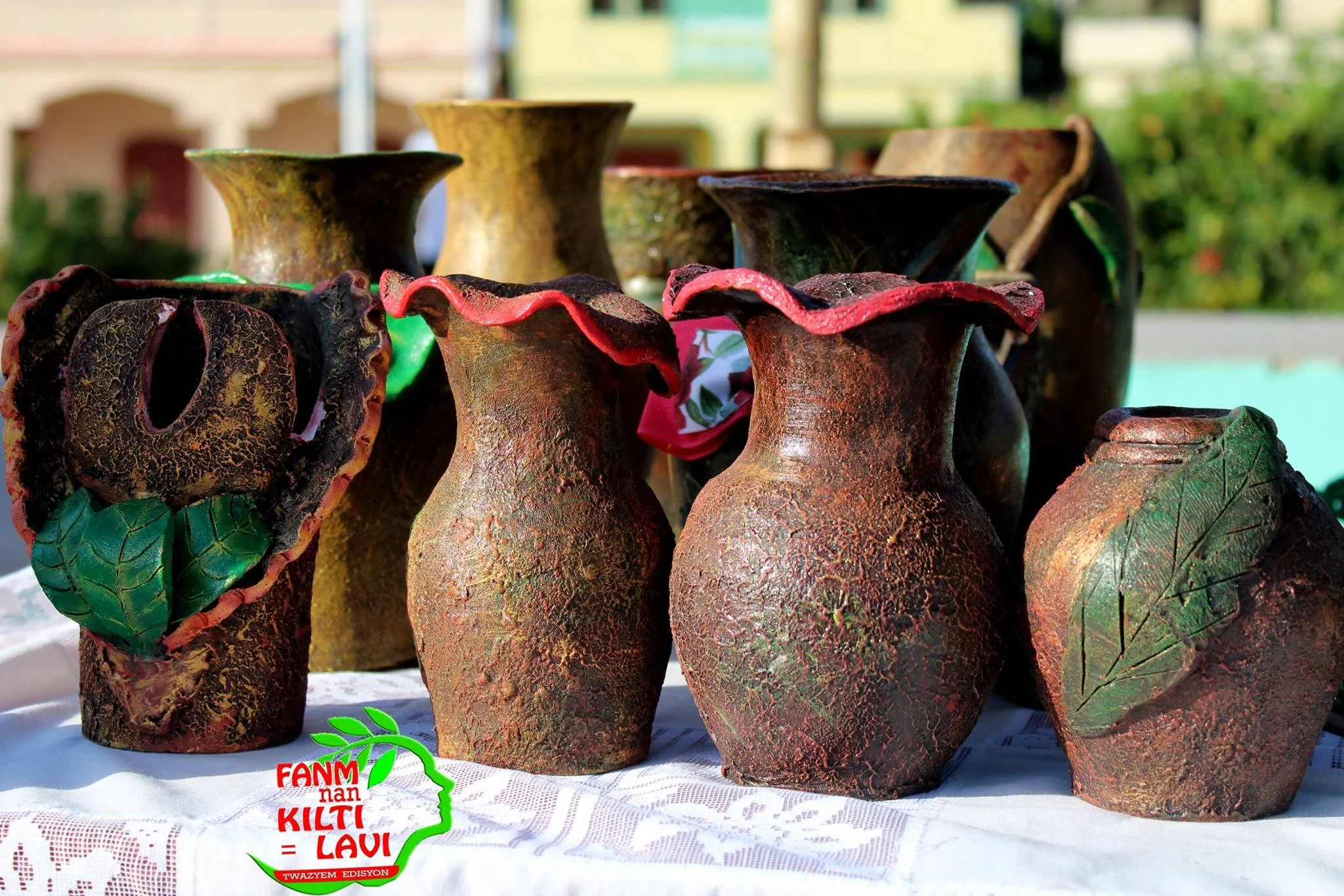 Vases en terre cuite artisanaux exposés sur une table, avec un fond flou de bâtiments et de végétation, représentant une scène de marché en plein air.