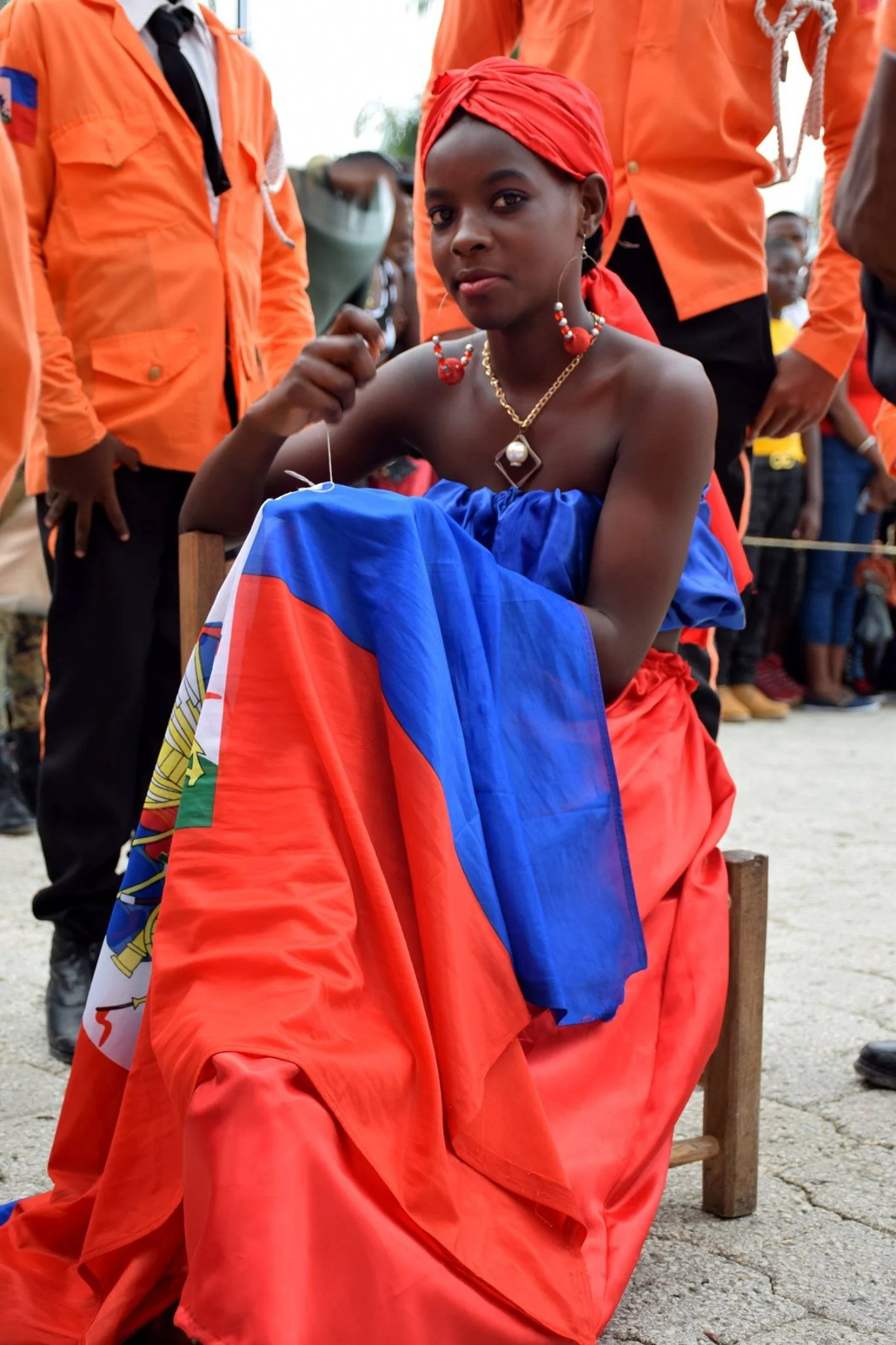 Une jeune femme vêtue d'une robe bleue, portant des bijoux, portant un foulard rouge sur la tête, assise sur une chaise en bois, entourée de personnes en vêtements orange.