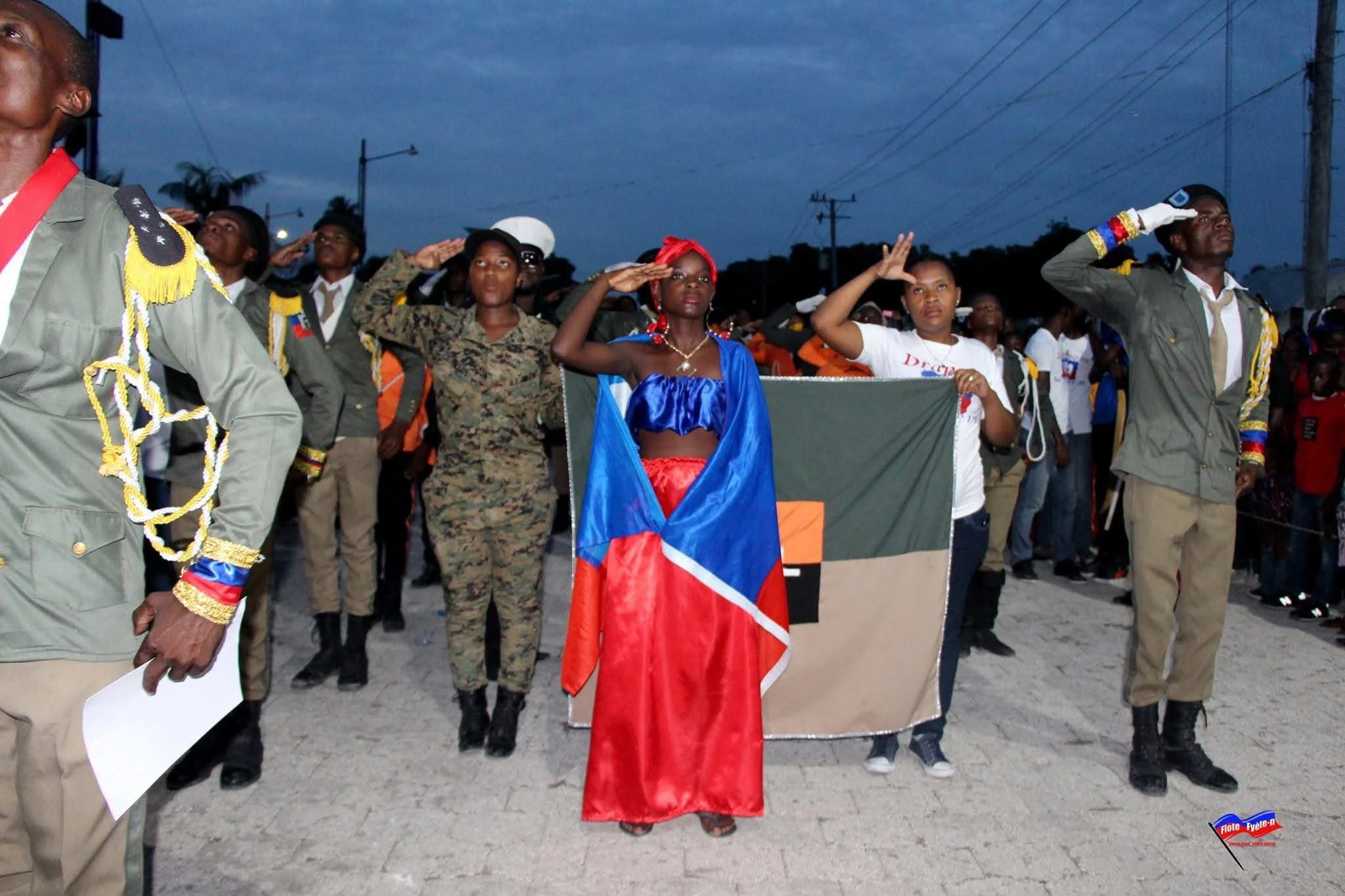 Groupe de personnes faisant un salut militaire lors d'une cérémonie en plein air, avec des drapeaux et des uniformes militaires, au crépuscule.