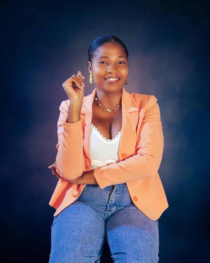 Jeune femme souriante portant un blazer orange, un haut blanc, des jeans bleus, avec des bijoux dorés, assise contre un fond sombre.
