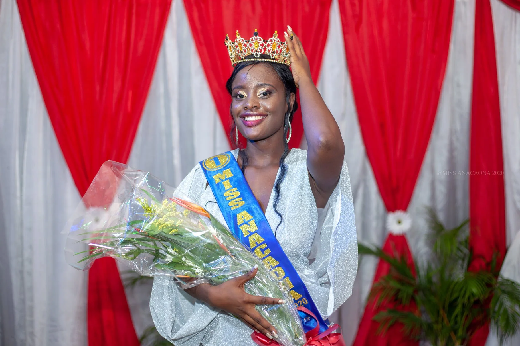 Femme couronnée portant une robe blanche, avec une écharpe bleue et un sash doré, tenant un bouquet de fleurs. Elle sourit et ajuste sa couronne, en arrière-plan un rideau rouge et blanc avec des plantes vertes.