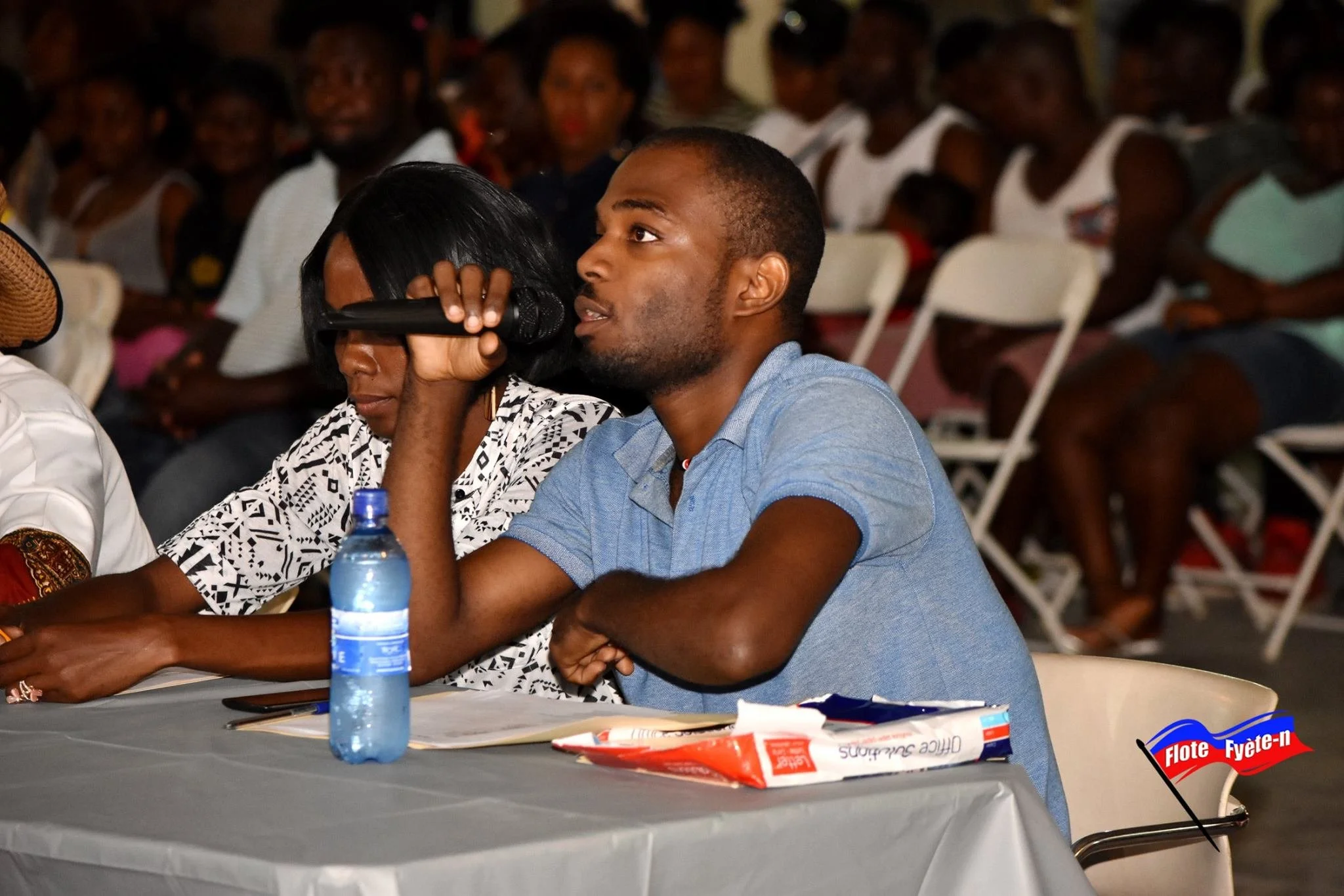 Un jeune homme assis à une table lors d'une fête, il regarde attentivement tandis qu'une femme à côté de lui utilise un microphone. Sur la table, il y a une bouteille d'eau, un paquet de chips et un téléphone. En bas à droite, un logo de fête indique