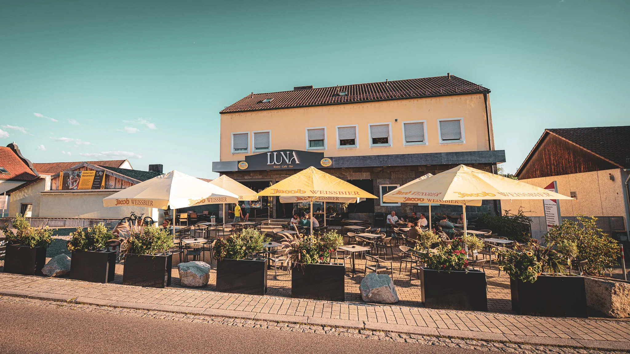Ein Restaurant mit dem Namen 'LUNA', Außenbereich mit Tischen und Sonnenschirmen, einige Gäste sitzen im Sommer, Gebäude mit gelben Wänden und roten Dachziegeln, blühende Pflanzen im Vordergrund.