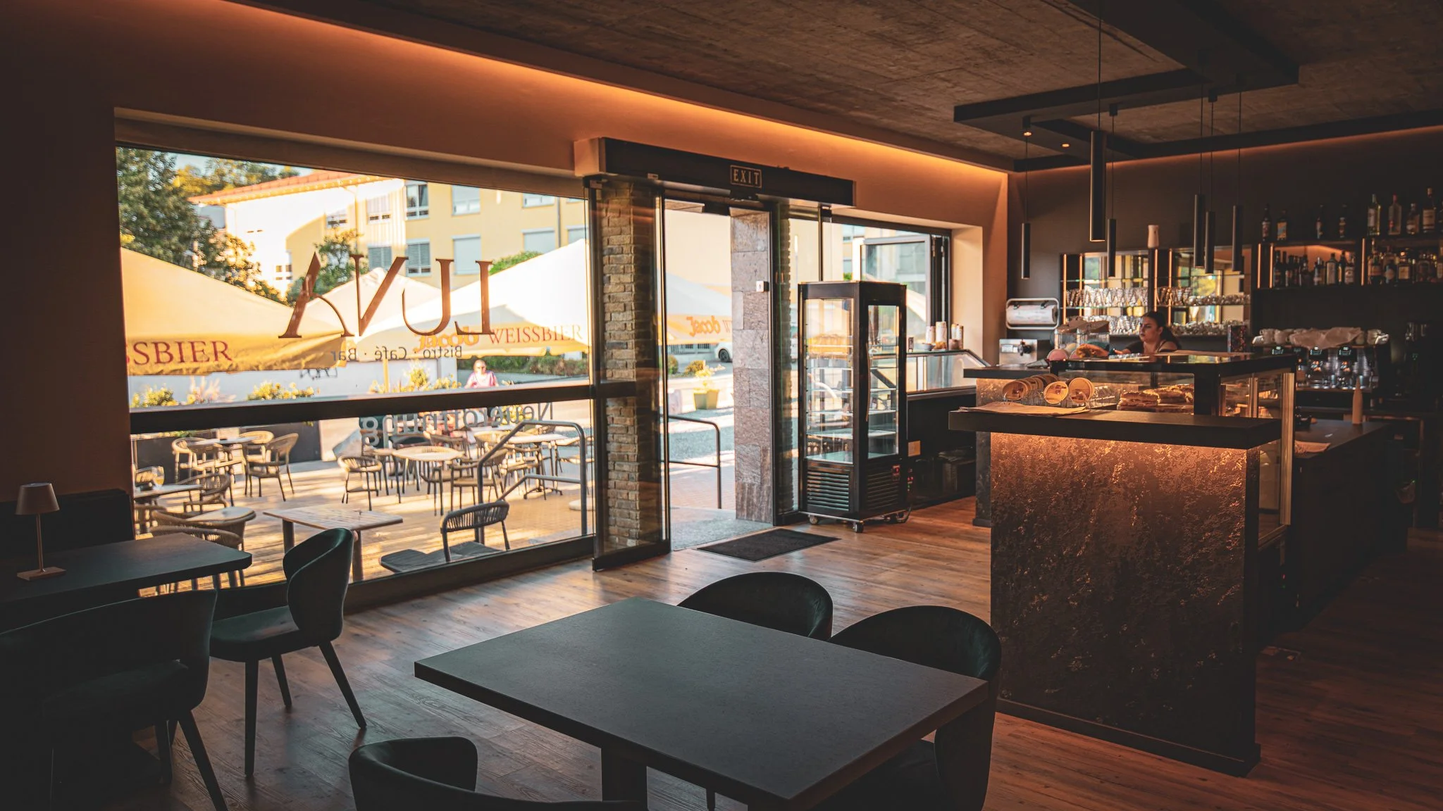 Innenansicht eines modernen Cafés mit großen Fenstern, die Tageslicht und Blick nach draußen auf eine Terrasse mit Tischen und Stühlen bieten. Im Hintergrund eine Bar mit Getränken und einem Barkeeper.