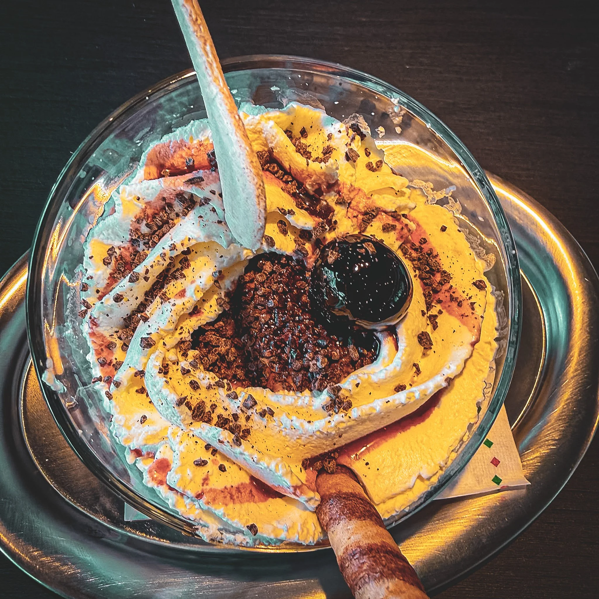 Schokoladeneis mit Bananen, Schokostreuseln, Kirschen und Vanilleeis in einer Glasschale mit Holzlöffel auf dunklem Tisch.
