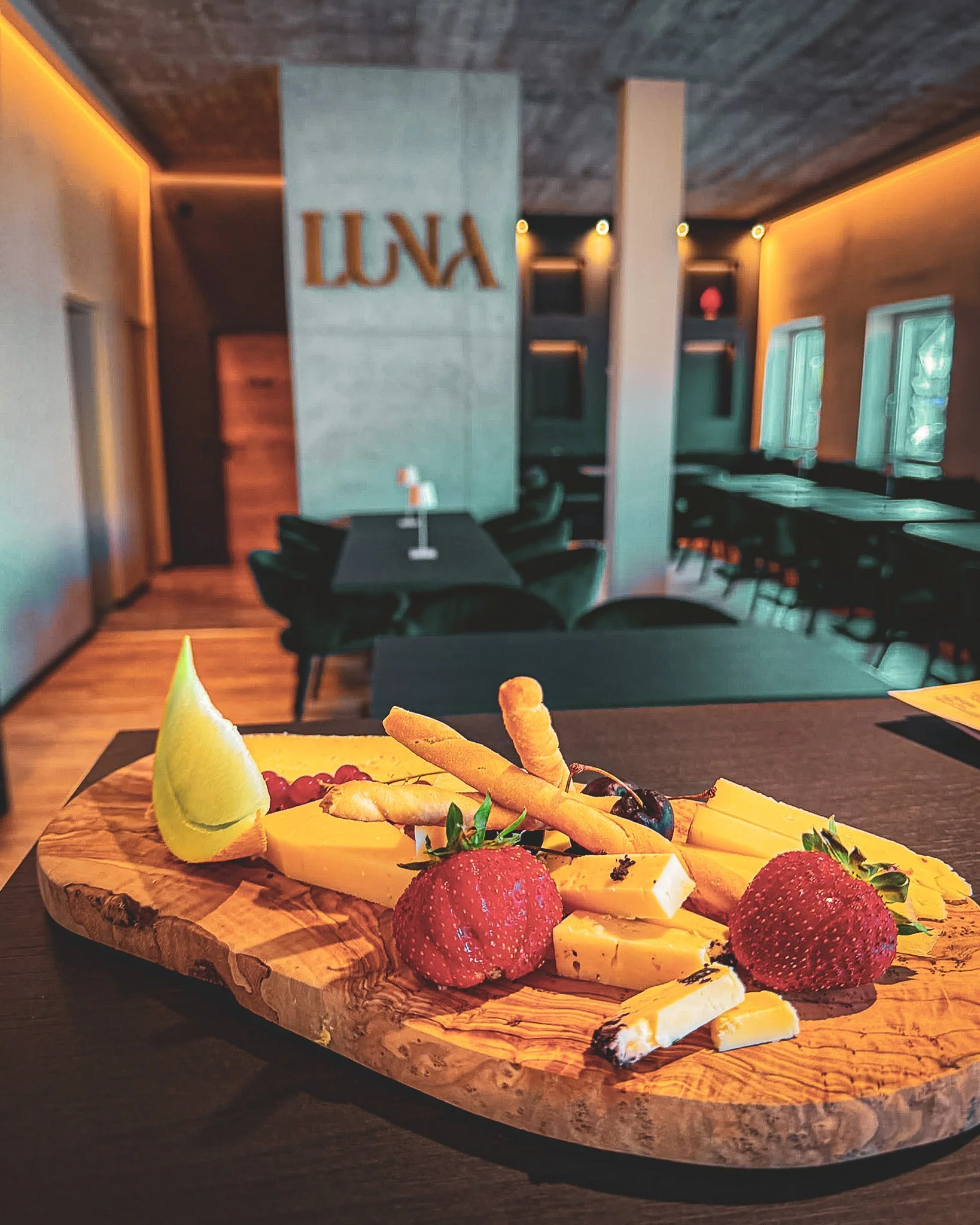 Käseplatte mit Erdbeeren, Weintrauben, Frischkäse, Schinken, Keksen, Melone, und mehreren Sorten Käse auf einem Holzbrett, serviert auf einem Tisch, im Hintergrund ein Restaurant mit Tisch und Stühlen, mit einer Wand mit der Beschriftung "LUNA" und d