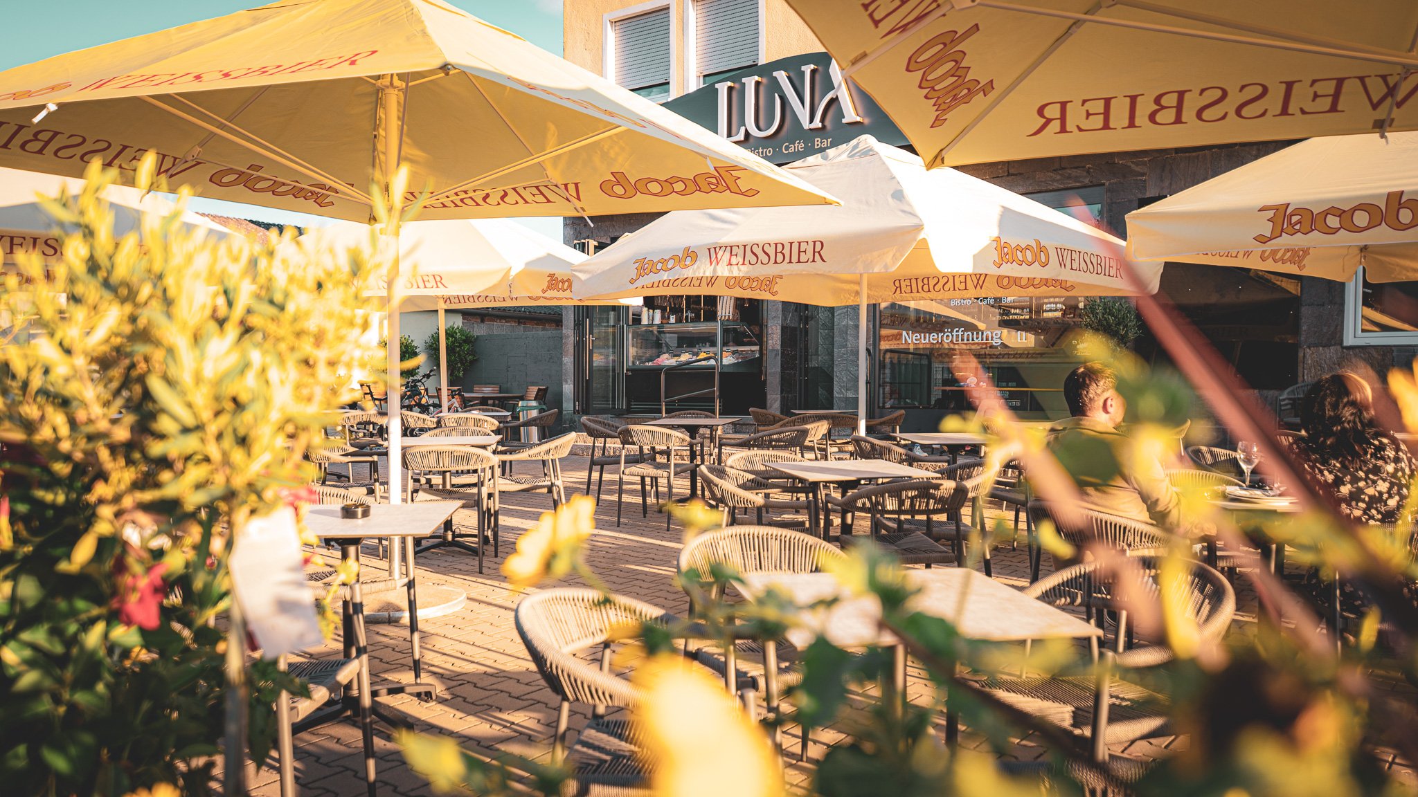 Ein leerer Außenbereich eines Cafés mit mehreren Stühlen und Tischen, große Sonnenschirme mit Werbetext, im Hintergrund eine Person bei Tisch, das Café heißt LUV. Es ist wahrscheinlich später Nachmittag, bei Sonnenuntergang.