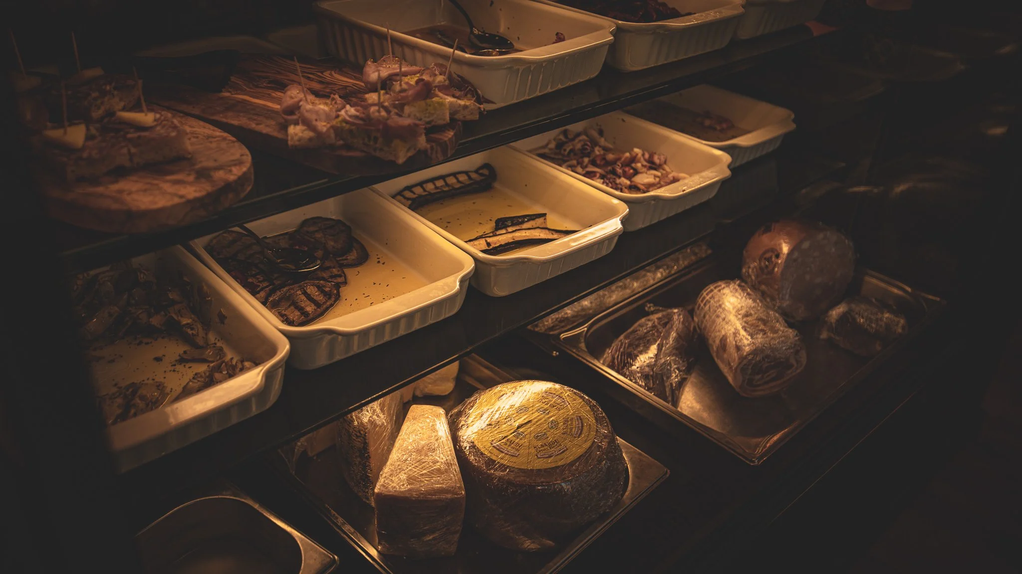 Verschiedene Speisen auf einem dunklen Buffet, einschließlich Fleisch, Käse, Aufschnitt und Vorspeisen in weißen Aufbewahrungsbehältern, bei schwachem Licht.