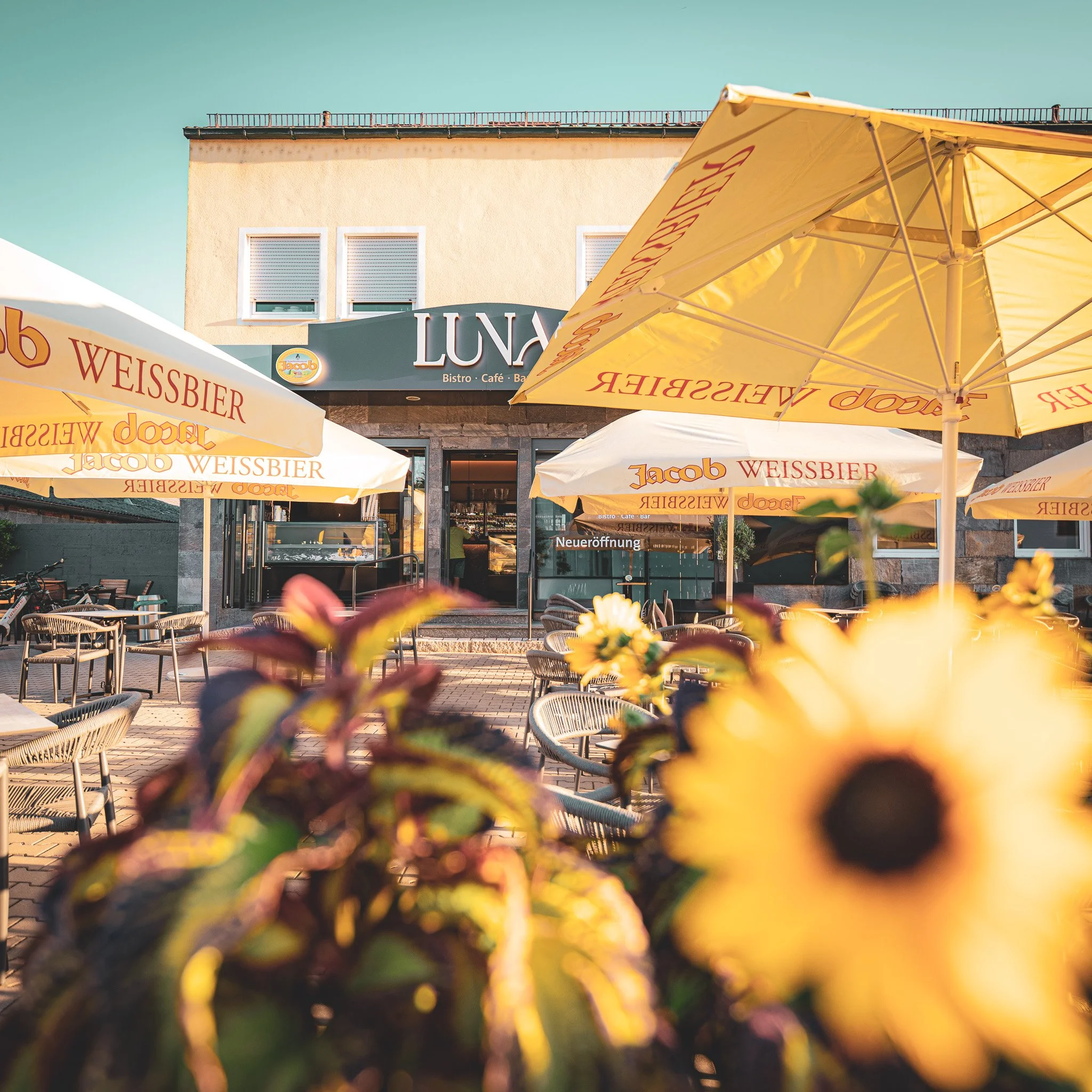 Außenbereich eines Cafés mit gelben Sonnenschirmen, die das Logo 'Jacob Weissbier' tragen, vor einem hellgelben Gebäude mit einem Restaurant- oder Café-Schild.