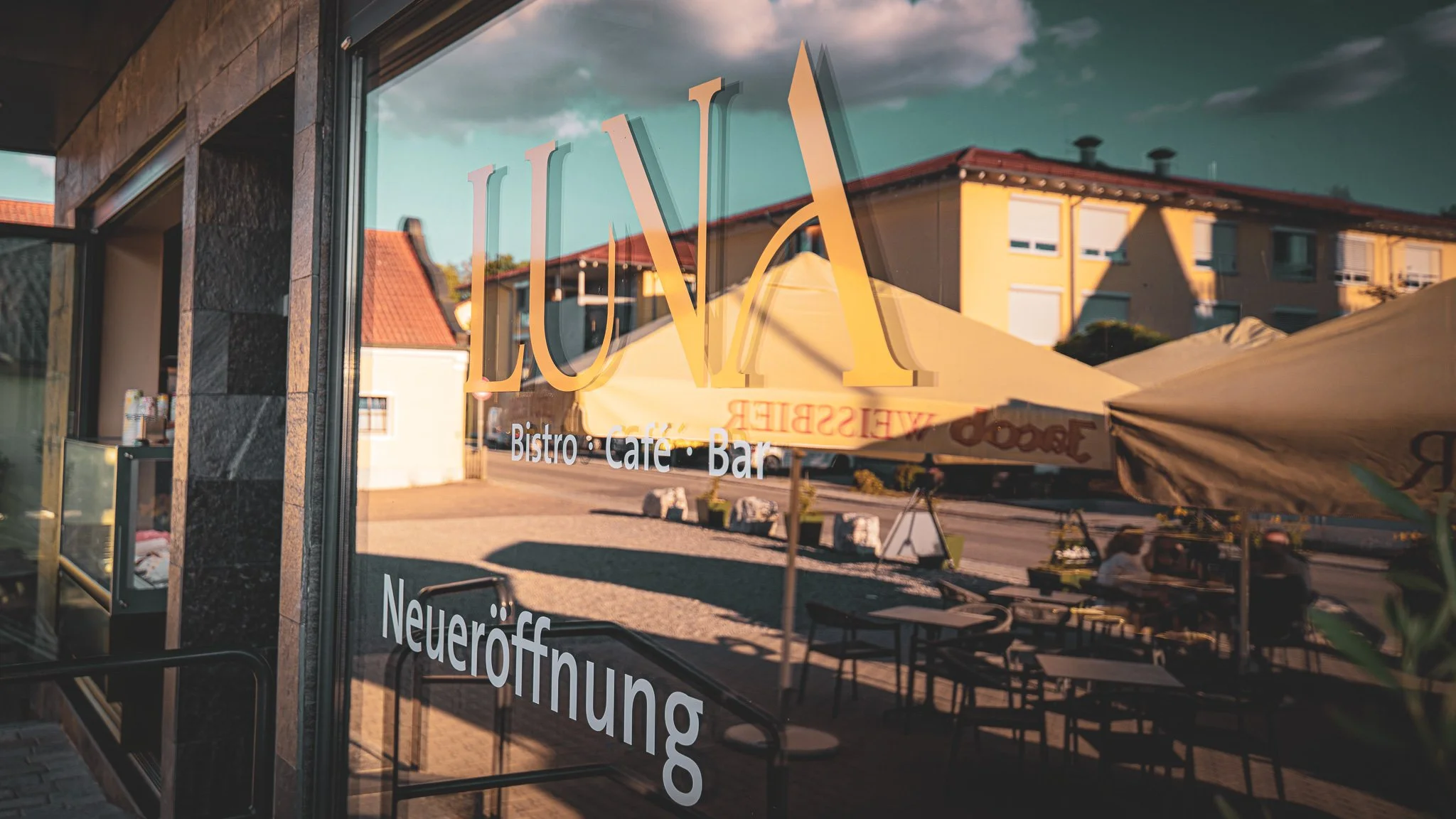 Fenster eines Cafés mit Schriftzug 'LUNA' und weiteren Angaben wie 'Bistro', 'Café', 'Bar'; draußen stehen Tische und Sonnenschirme, im Hintergrund Wohnhäuser bei Sonnenuntergang.