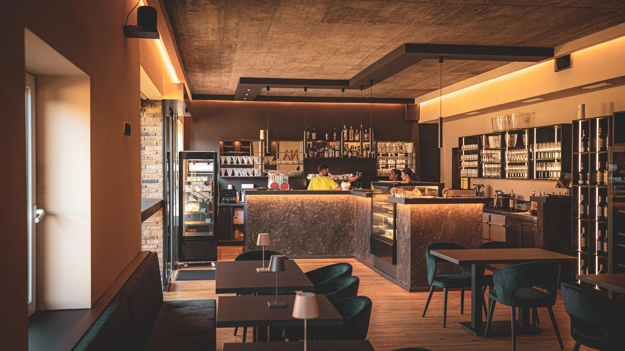 Innenaufnahme einer Bar oder eines Cafés mit Sitzgelegenheiten, dunkler Theke und ein paar Leuten, die sich unterhalten, moderne Beleuchtung und freundliche Atmosphäre.