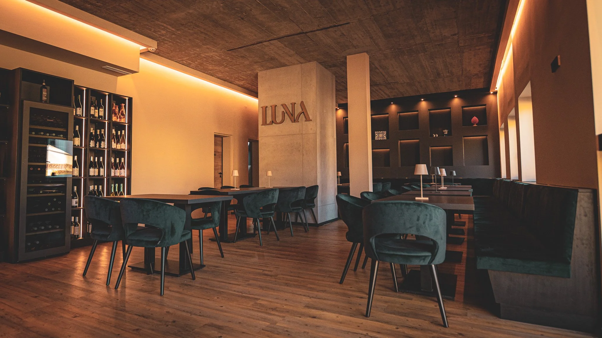 Innenaufnahme eines modernen, eleganten Restaurants mit dunklen Stühlen, Holzboden, Regal mit Weinflaschen und dem Schriftzug 'LUNA' an einer Wand.