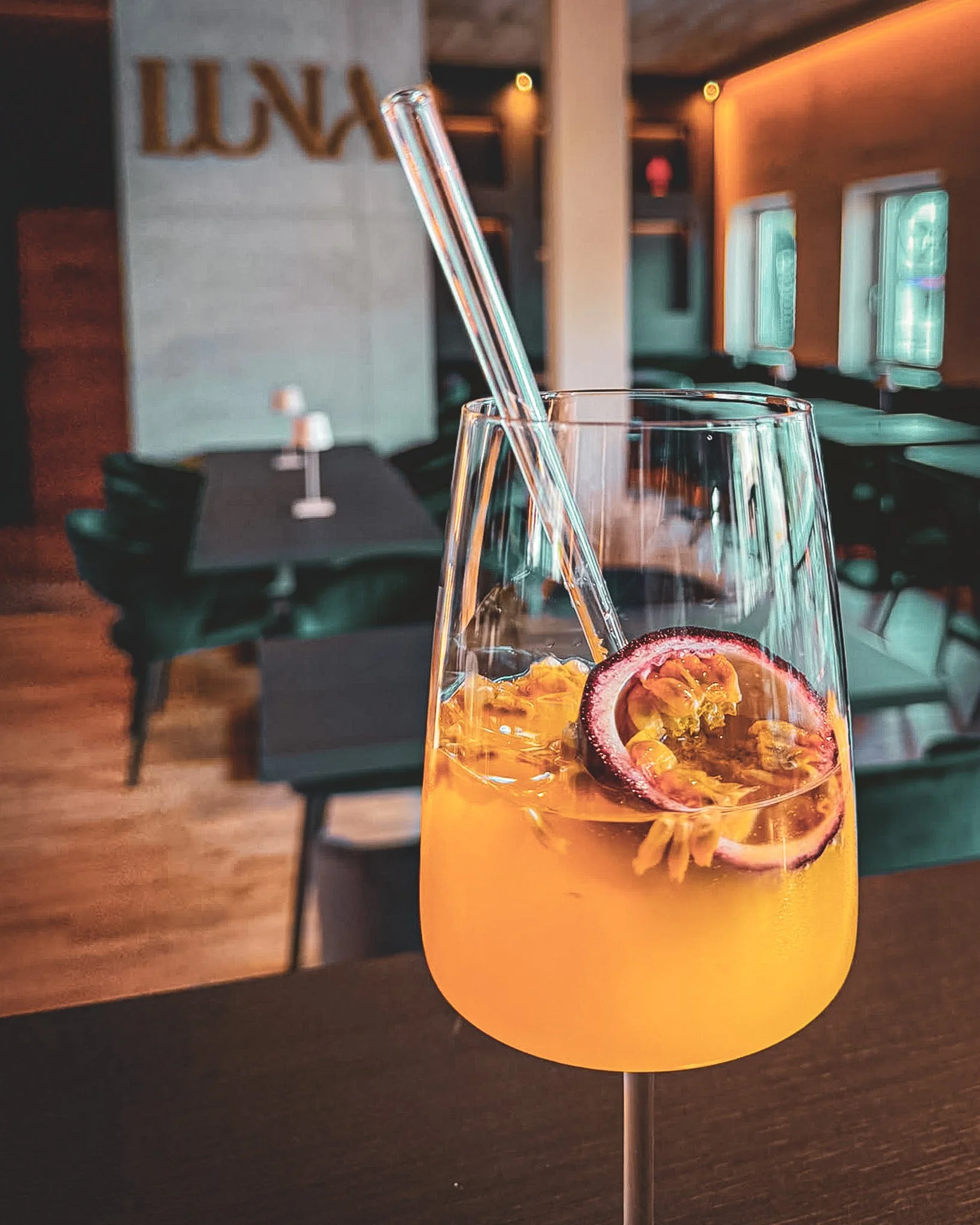 Ein Glas mit einem Getränk, das Passionsfrucht- und Orangenstücke enthält. Im Hintergrund sind Tische und Stühle in einem Café oder Restaurant sichtbar.