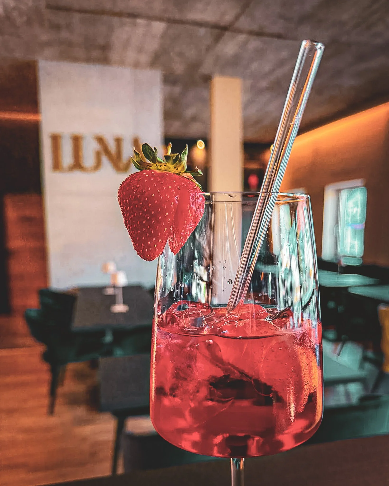Ein Glas mit rotem Getränk und Eiswürfeln, garnierte mit einer Erdbeere und einem Trinkhalm, im Hintergrund eine Bar oder ein Restaurant. 