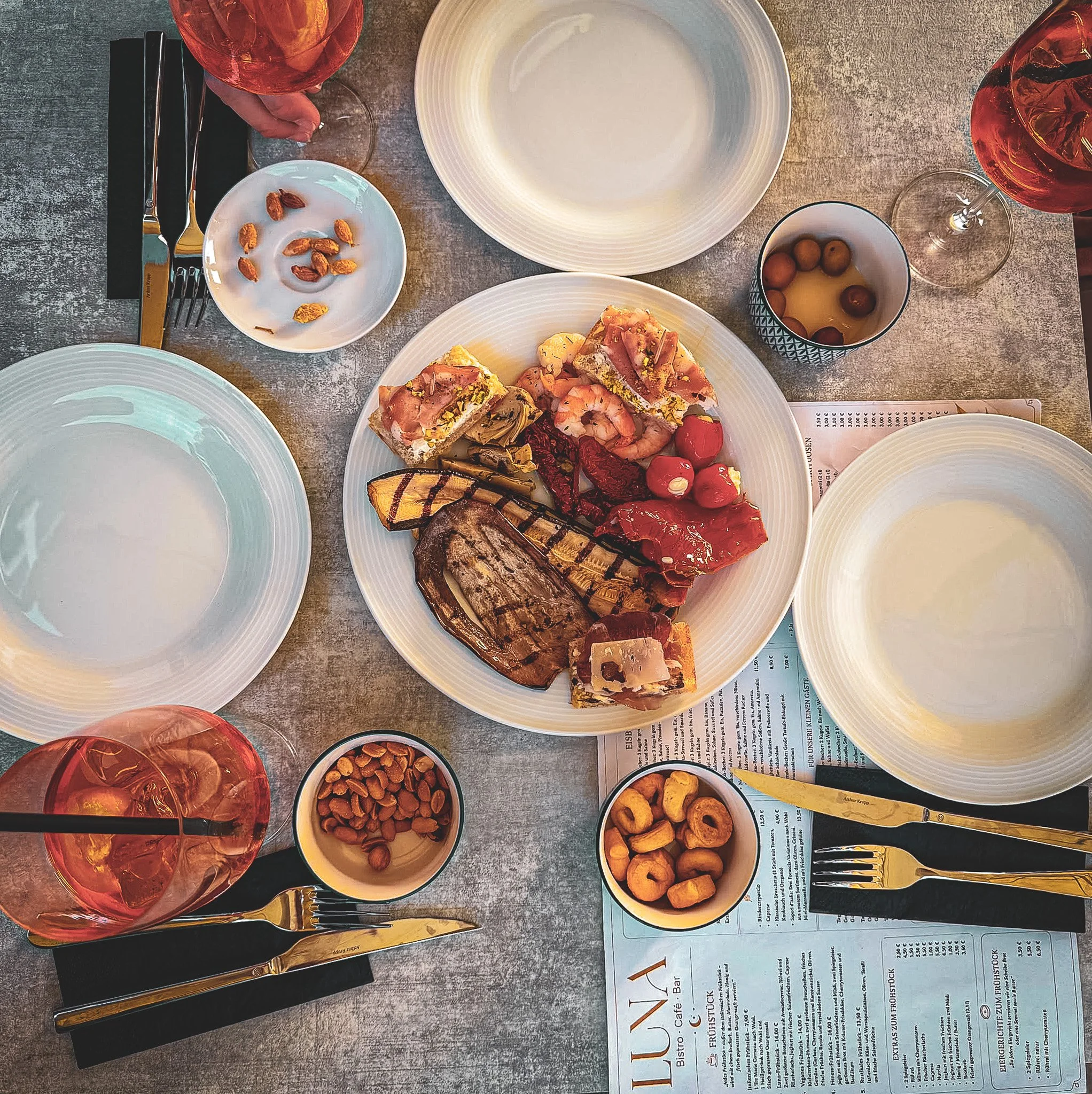 Ein gedeckter Esstisch mit leeren weißen Tellern, mehreren roten Wein- und Wasserweingläsern, diversen kleinen Schalen mit Snacks, einer großen Schale mit belegten Pizzastücken, einem Stück gegrilltem Fleisch, einem Salat und einem Stück Kuchen, sowi