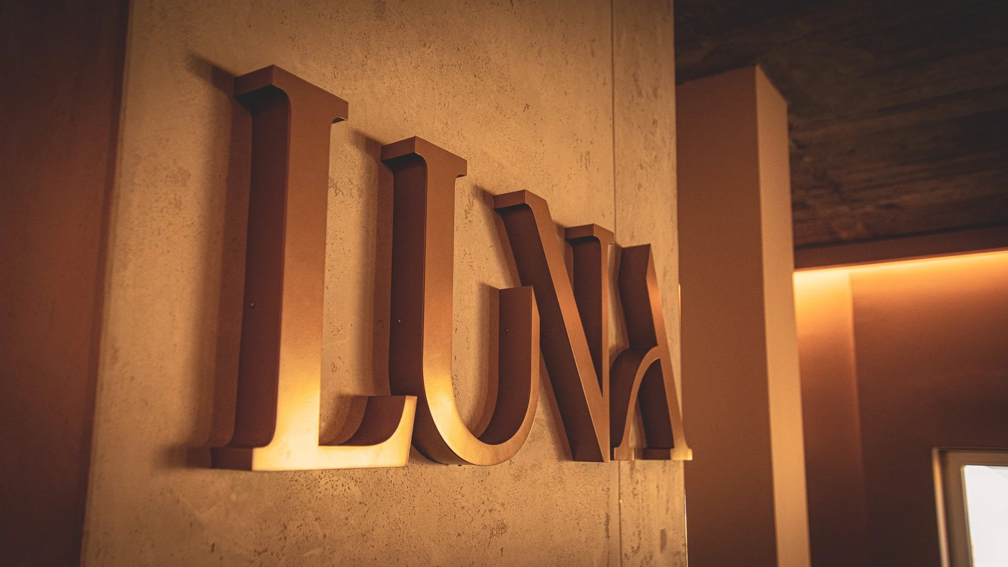 Nahaufnahme eines Wanddekors mit großen, dreidimensionalen Buchstaben, die an einer Wand angebracht sind, in warmem Licht. Im Hintergrund eine dunkle Decke und eine Wand mit einer abgedeckten Lichtquelle.