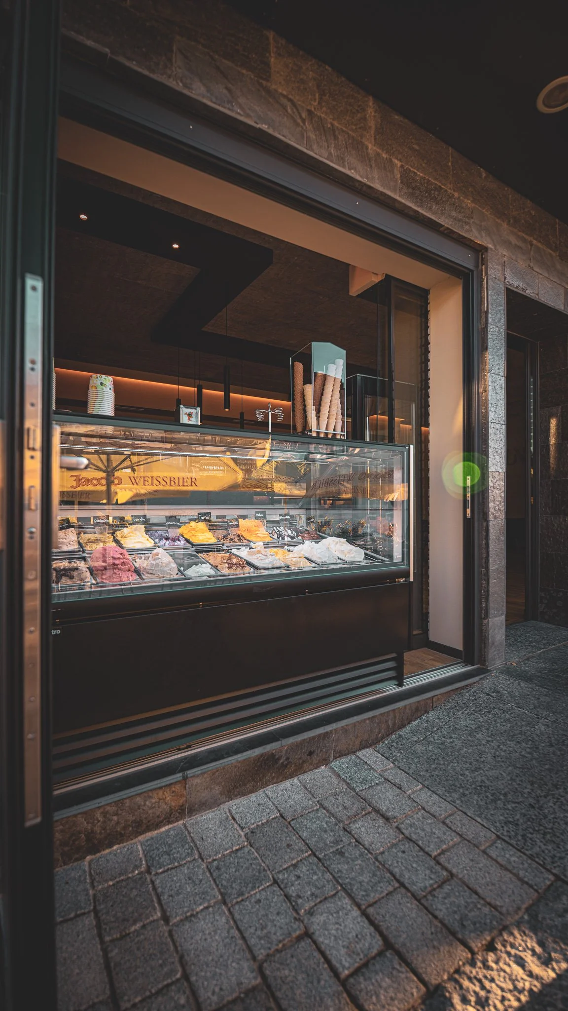 Eisverkaufsstand mit verschiedenen Eissorten in der Vitrine, draußen vor einem Gebäude mit Steinmauer und Pflastersteinen auf dem Gehweg.