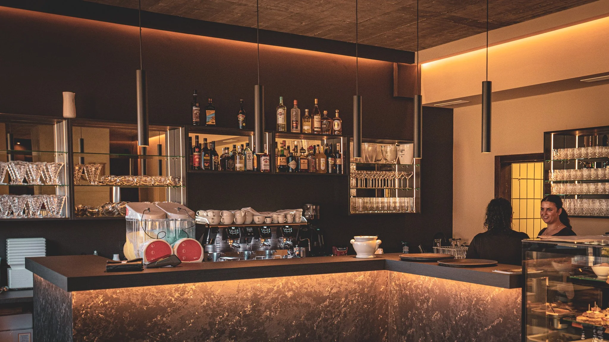 Innenansicht einer Bar mit dunklem Design, mehreren Flaschen alkoholischer Getränke auf Regalen, Gläsern, Tassen und Kaffeemaschinen auf der Theke, und zwei Frauen im Gespräch.