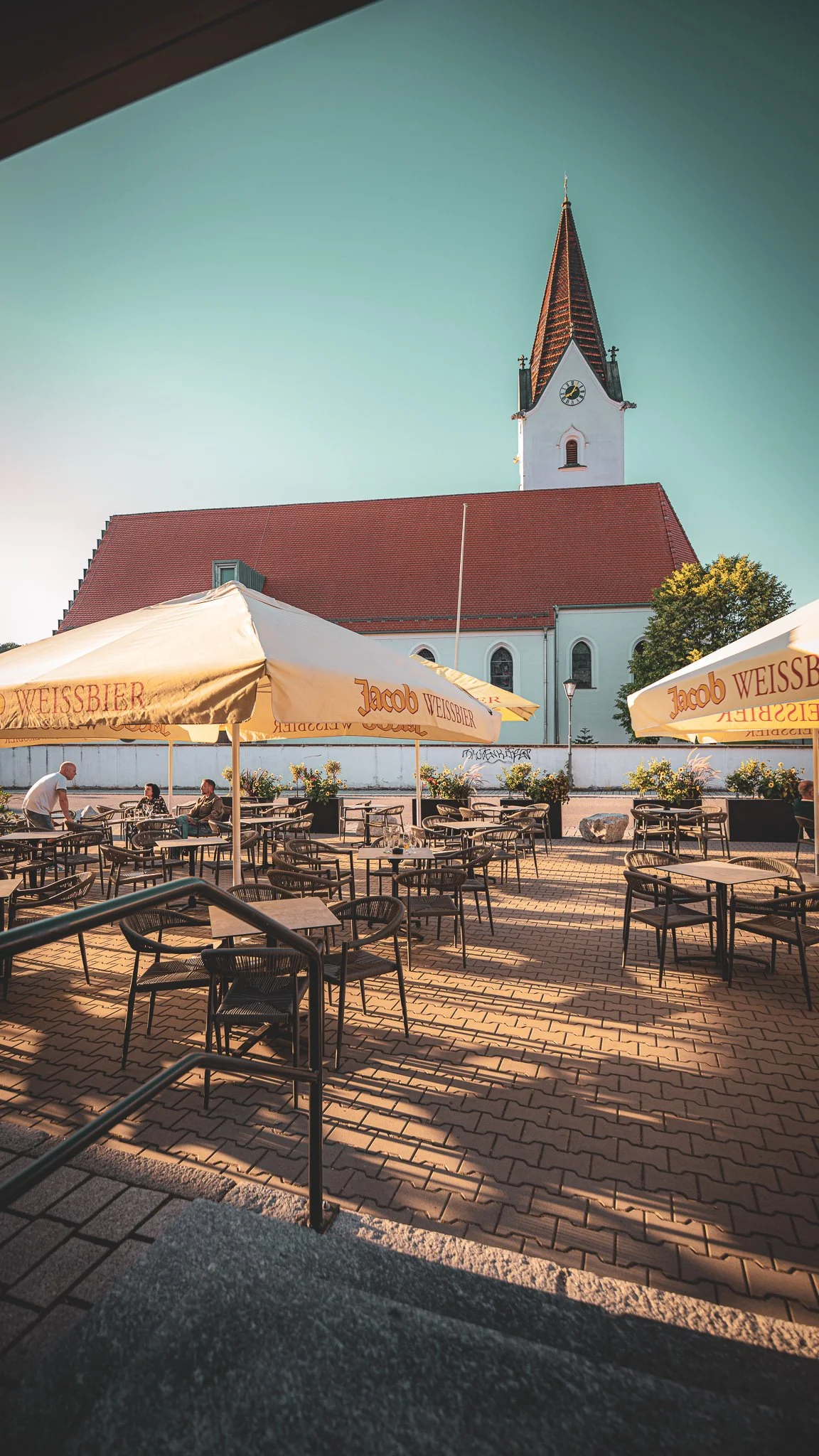 Ein leerer Außenbereich mit Tischen und Stühlen, Sonnenschirmen, vor einer weißen Kirche mit rotem Dach und Kirchturm, bei Sonnenuntergang.