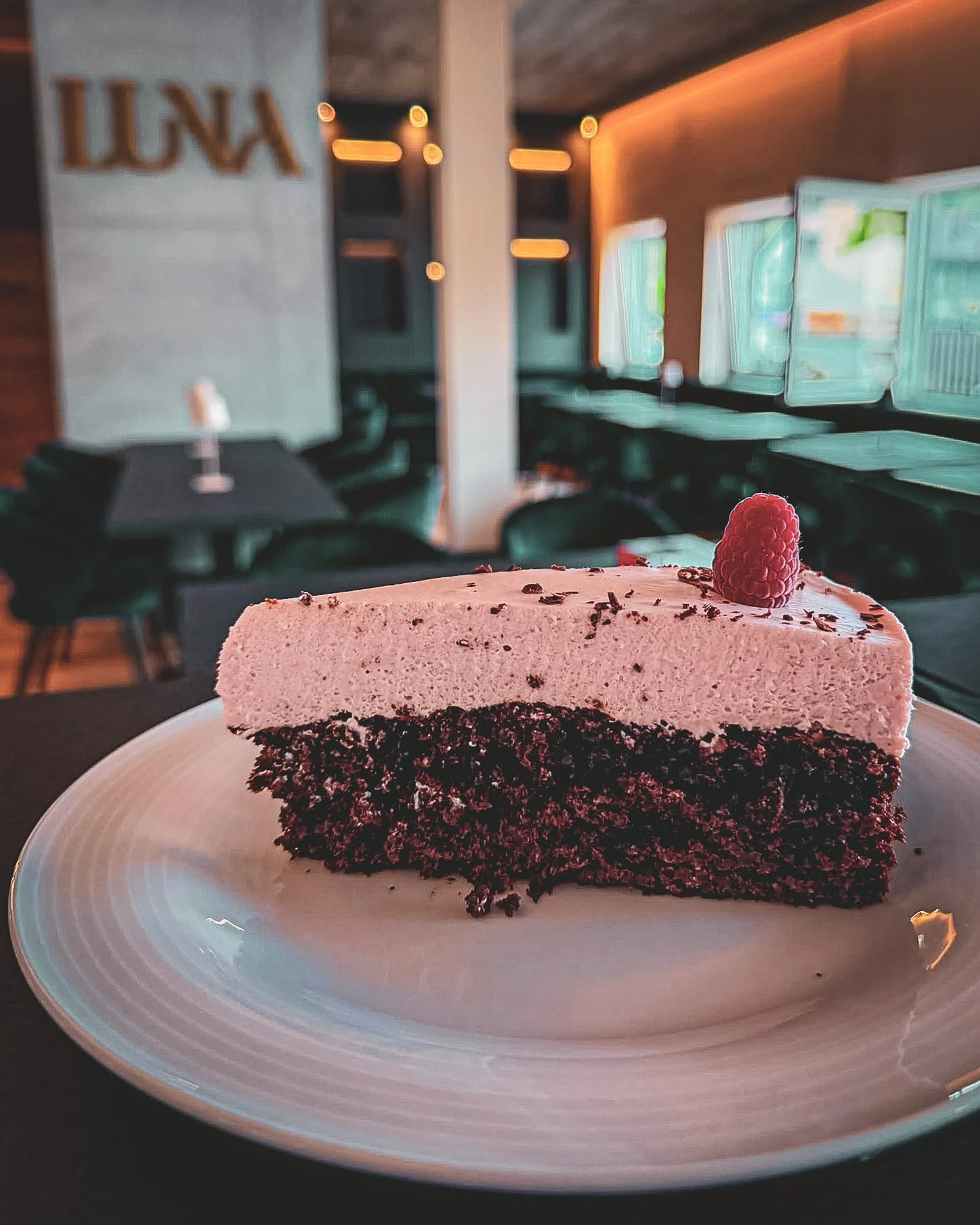 Ein Stück Schokoladenkuchen mit rosa Schaum und Himbeere auf einem weißen Teller, im Hintergrund ein Restaurant mit dunklen Stühlen und großen Fenstern.