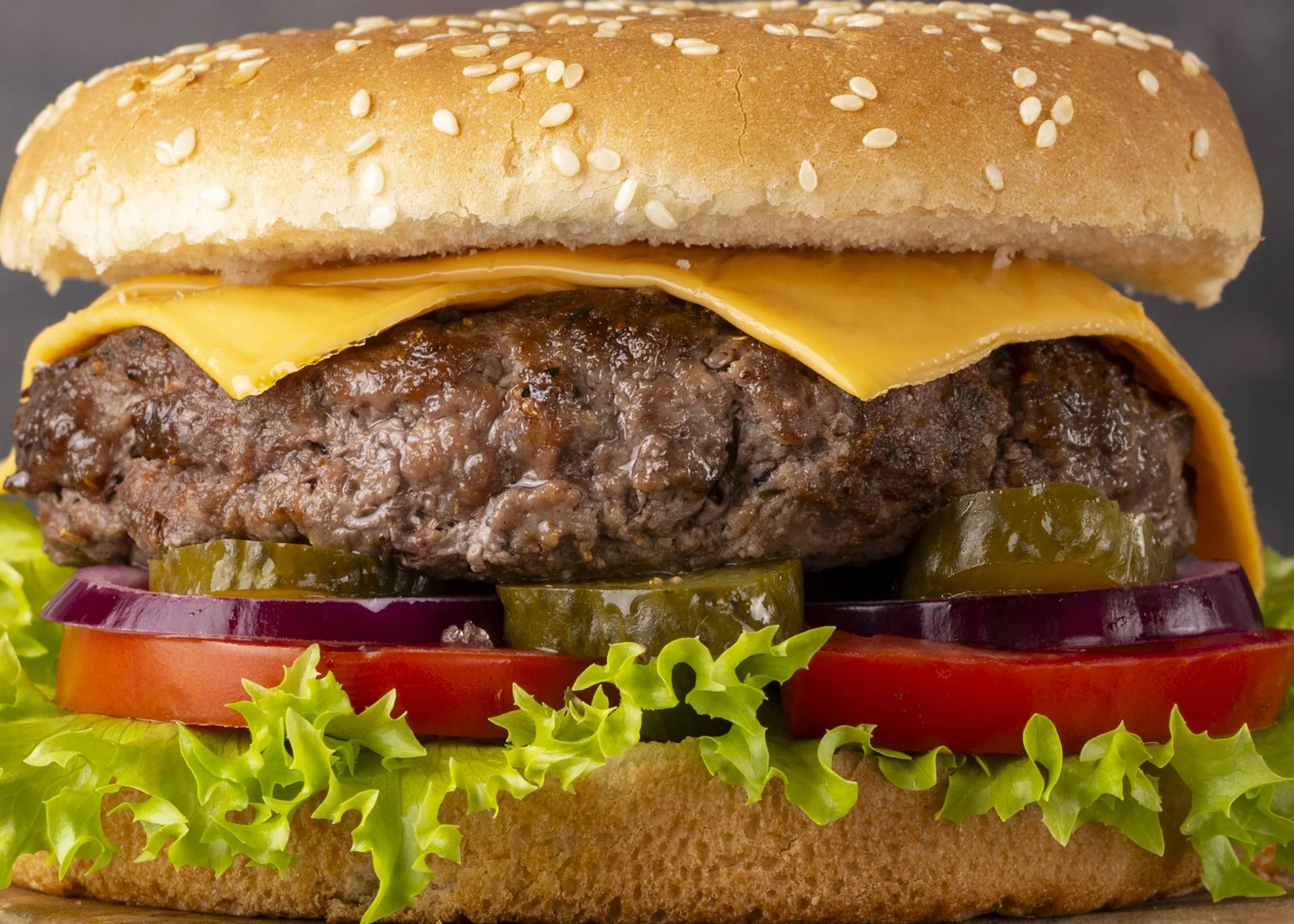 Close-up of a cheeseburger with a sesame seed bun, cheddar cheese, hamburger patty, tomato slices, pickles, red onion, lettuce, and other vegetables.