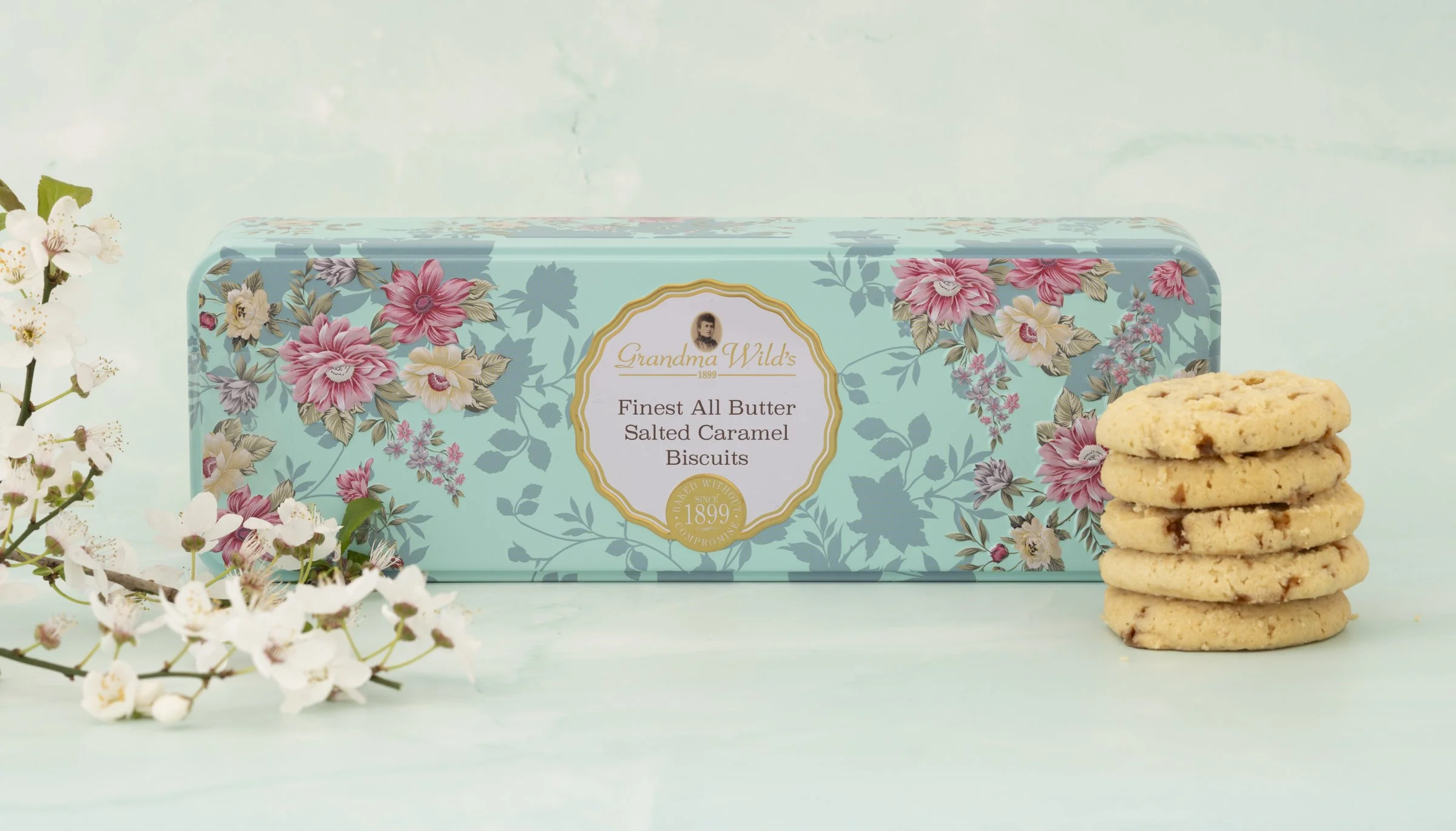 A box of Grandma Wild's salted caramel biscuits with floral packaging, next to a stack of four biscuits and some white flowers on a light blue background.