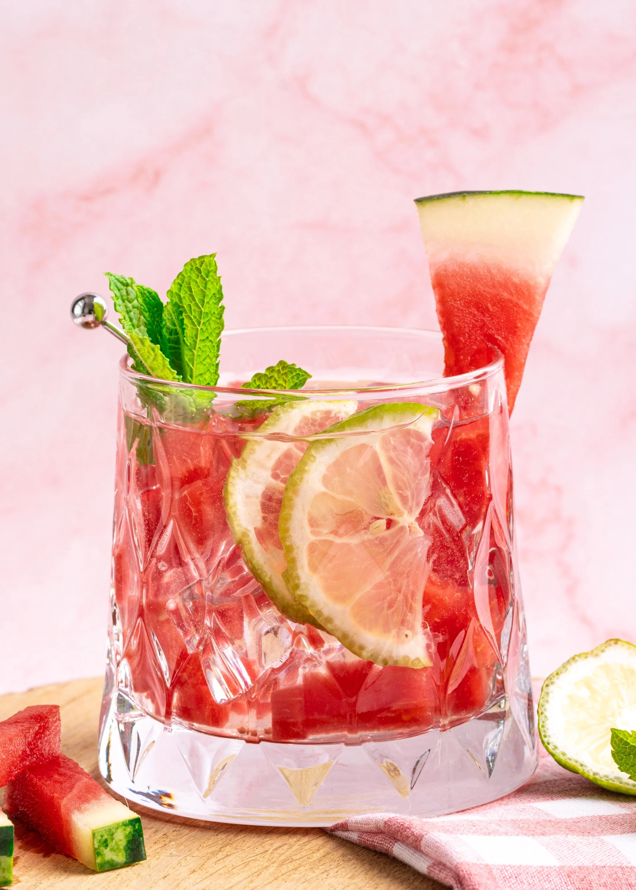 A glass of pink watermelon lemonade garnished with lemon slices, fresh mint, and a watermelon wedge on a pink background.