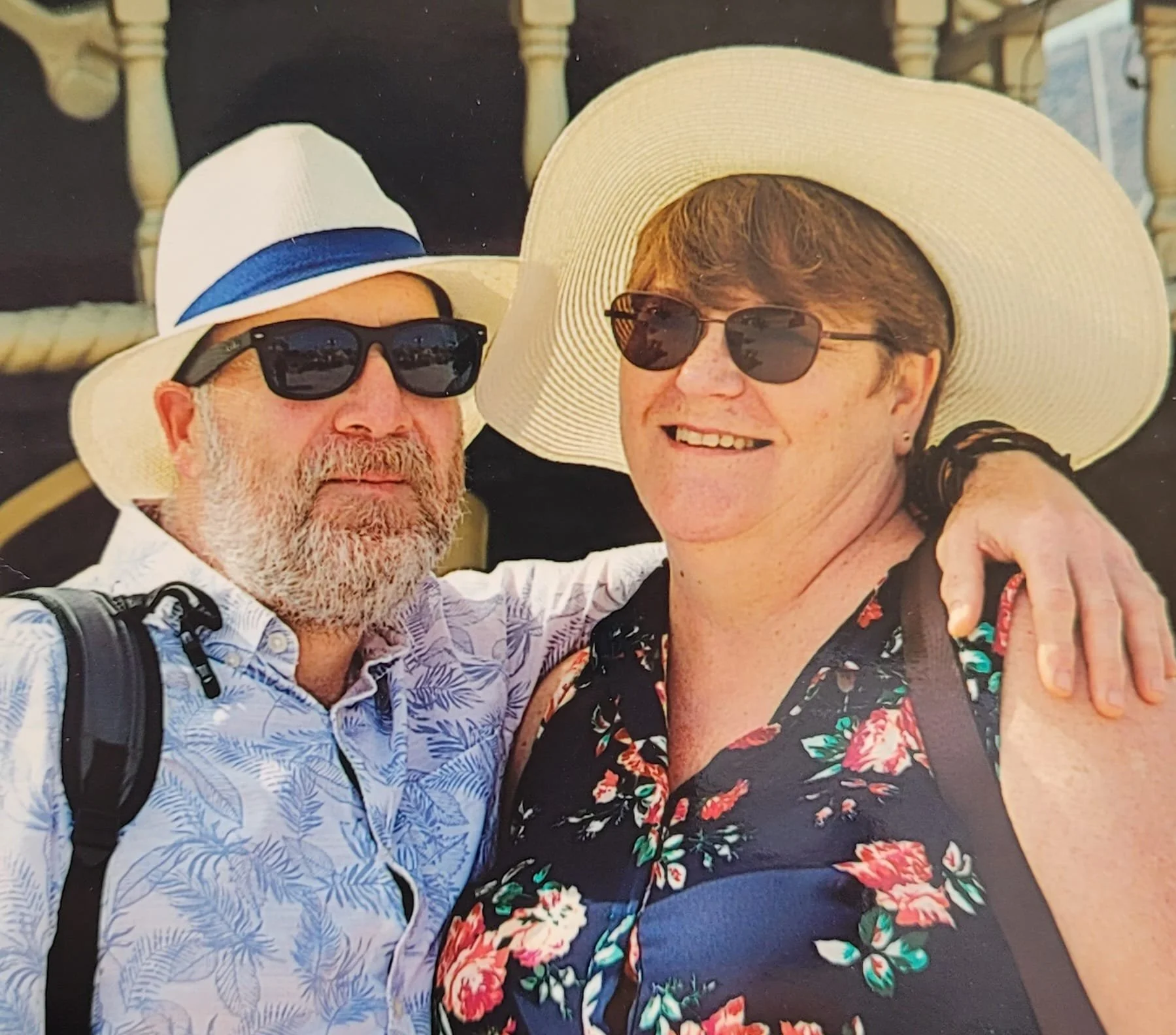 A smiling man with a beard and sunglasses, wearing a white hat with a blue band and a patterned shirt, standing next to a woman with short hair, glasses, a wide-brimmed hat, and a floral shirt. They are outdoors with a wooden structure in the background.