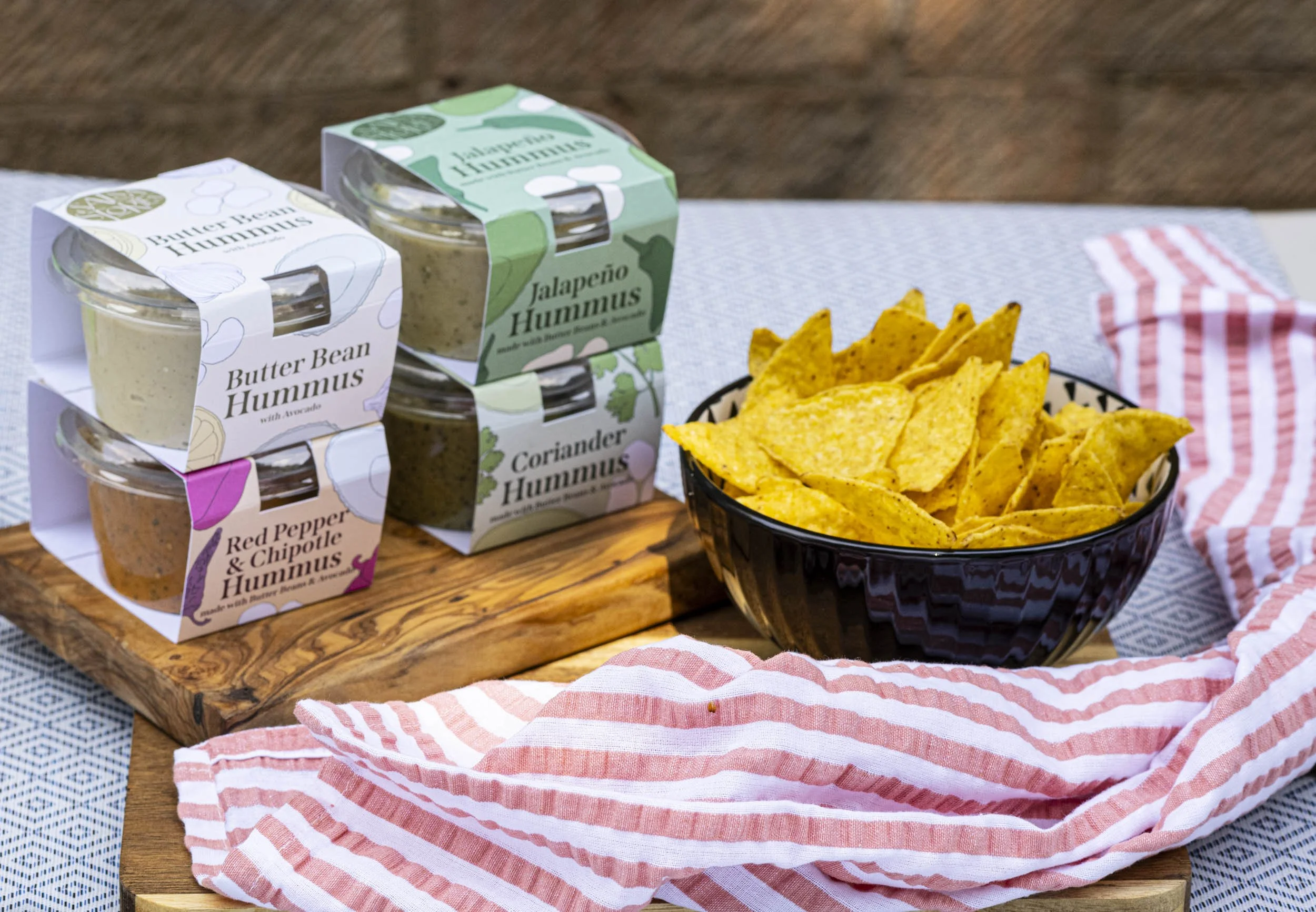 Variety of hummus containers with different flavors, a bowl of tortilla chips, and a red and white striped cloth on a wooden board.