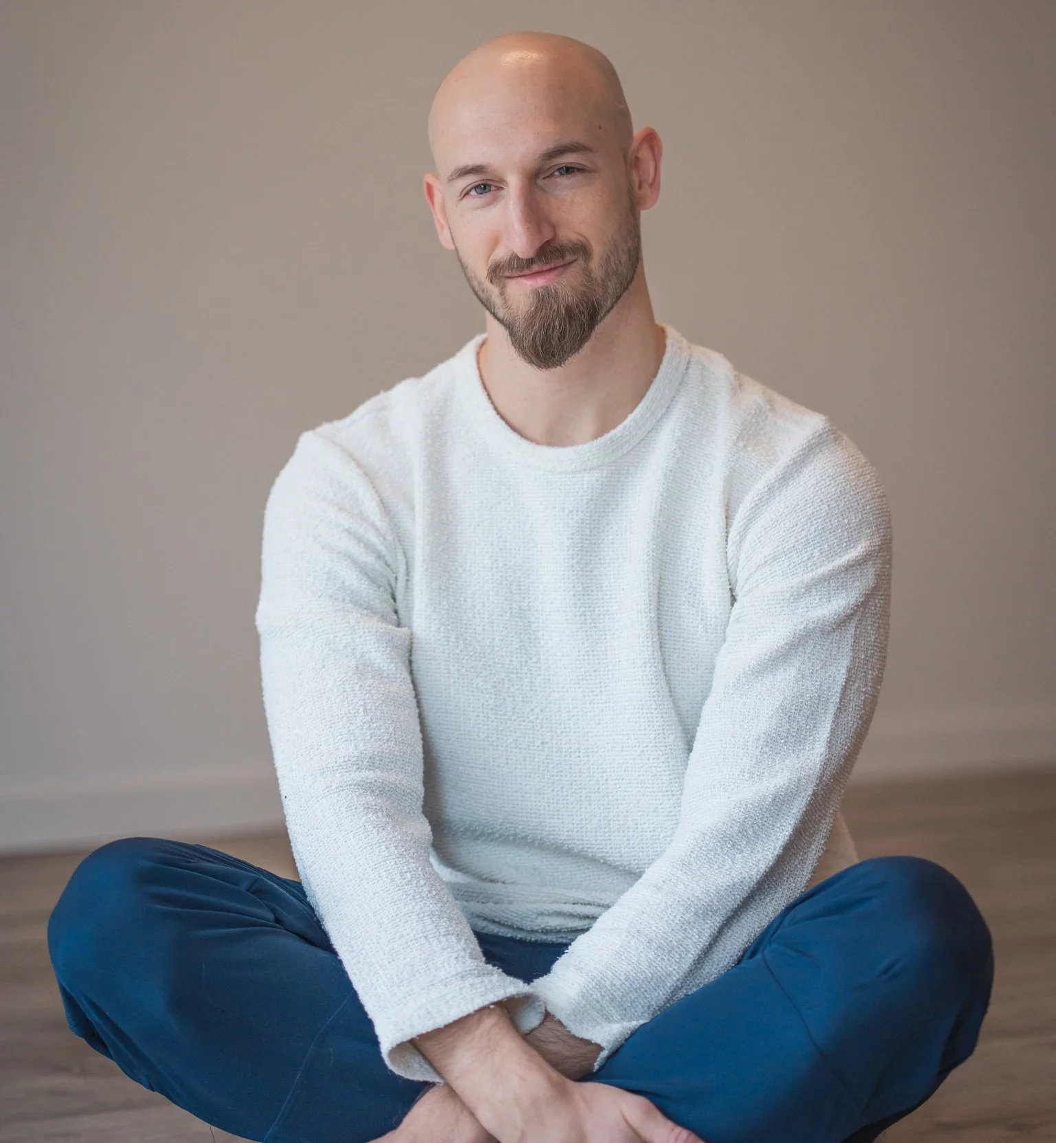 Adrian sitting cross-legged on the floor, wearing a white sweater and blue pants, smiling gently against a plain gray background.