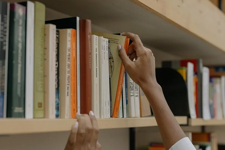 A person is reaching for a book on a wooden bookshelf, with their hand grasping the top edge of the book while their other hand holds the shelf to steady themselves.
